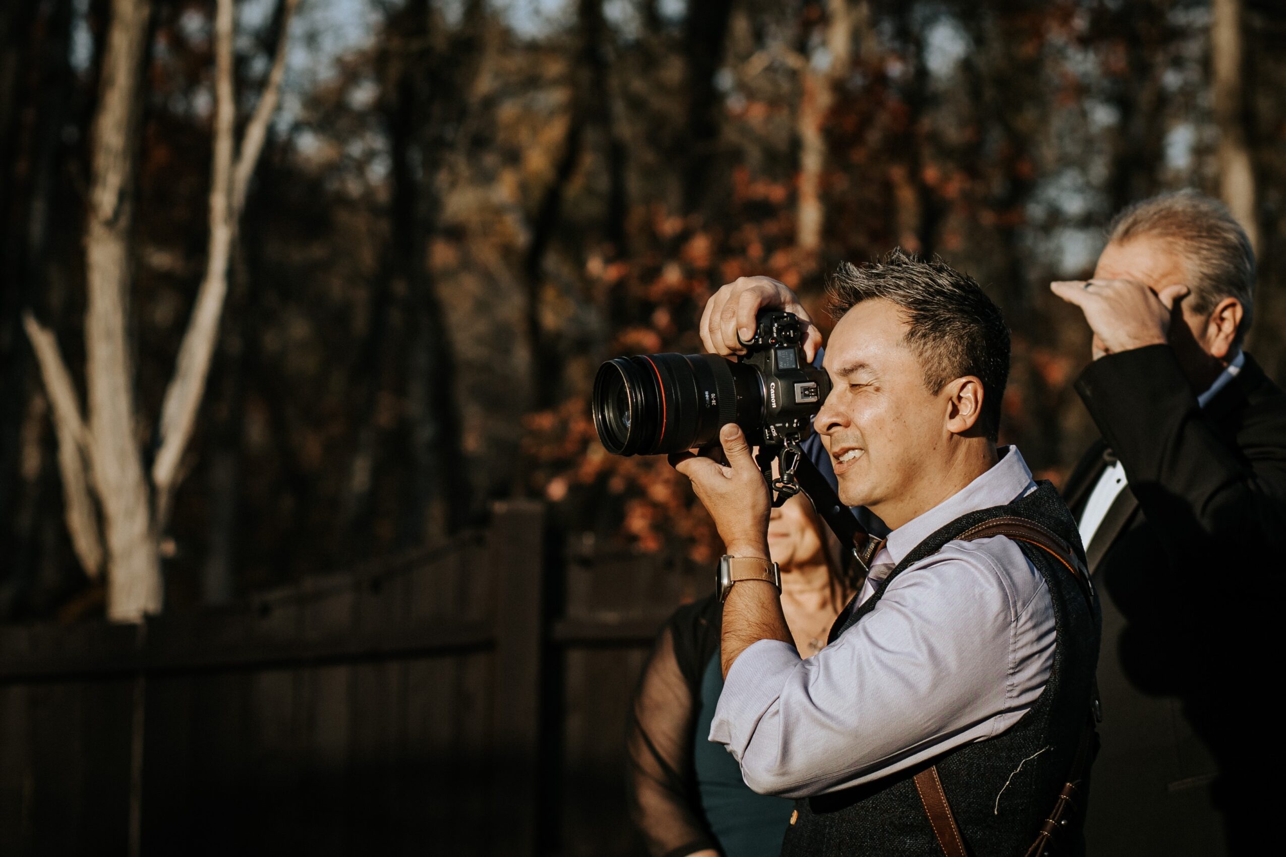 Two men outdoors, one taking a photo with a camera, trees with autumn leaves in background