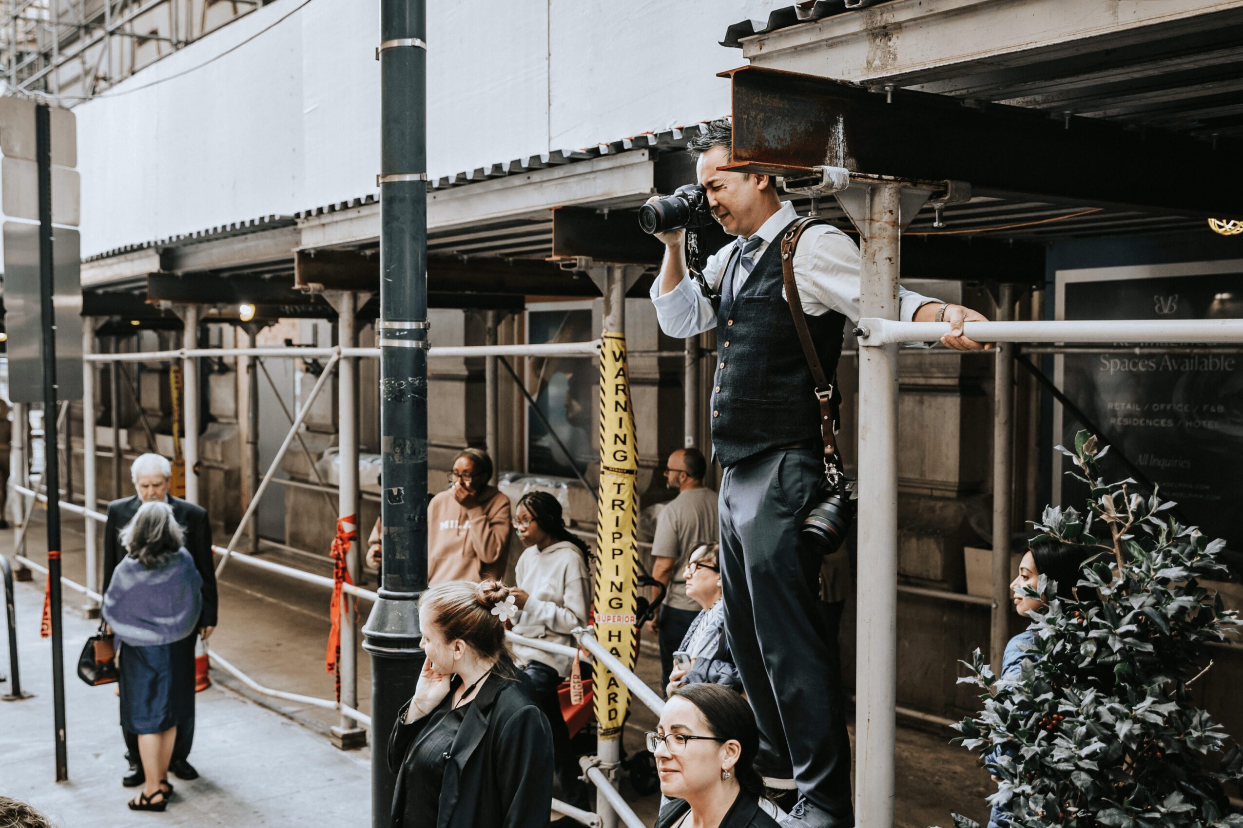 Photographer standing on a platform taking pictures on a city street with pedestrians and scaffolding nearby.
