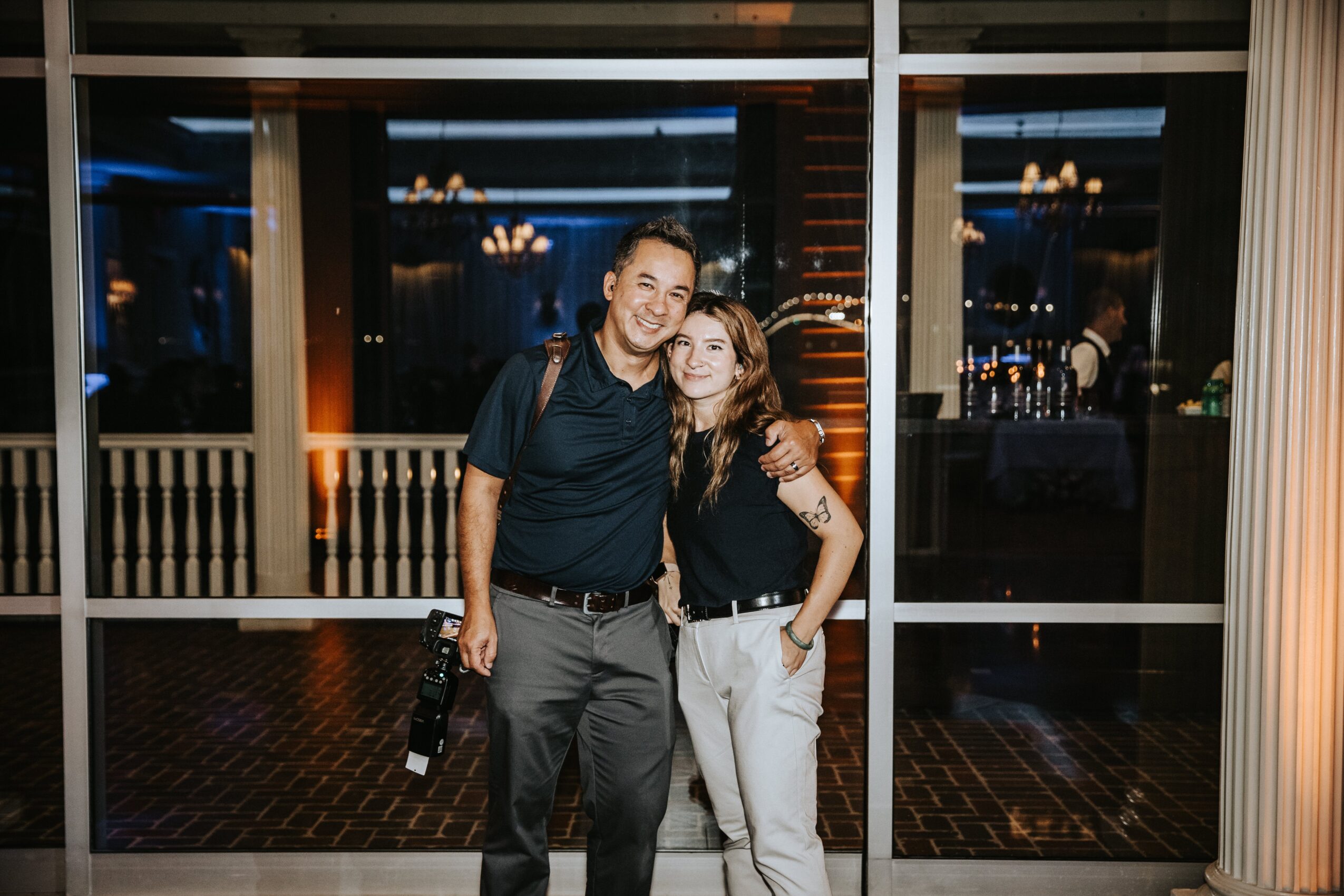 Two people standing indoors, smiling, with large windows and reflections behind them.