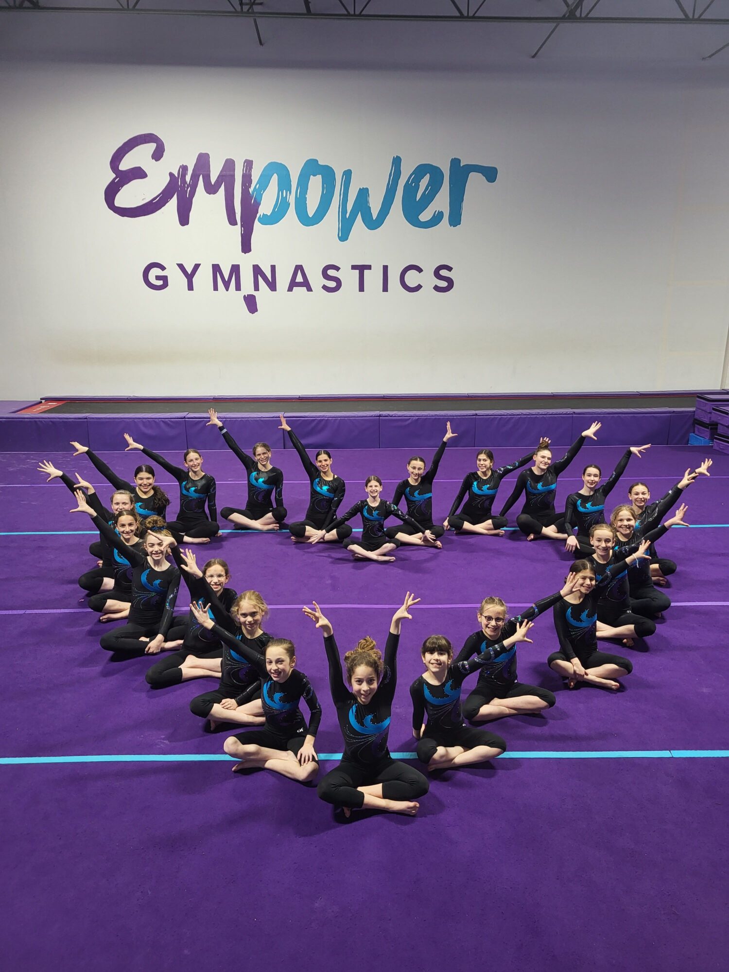 Group of gymnasts in black and blue uniforms performing a synchronized routine on purple mats, with a gymnasium wall in the background.