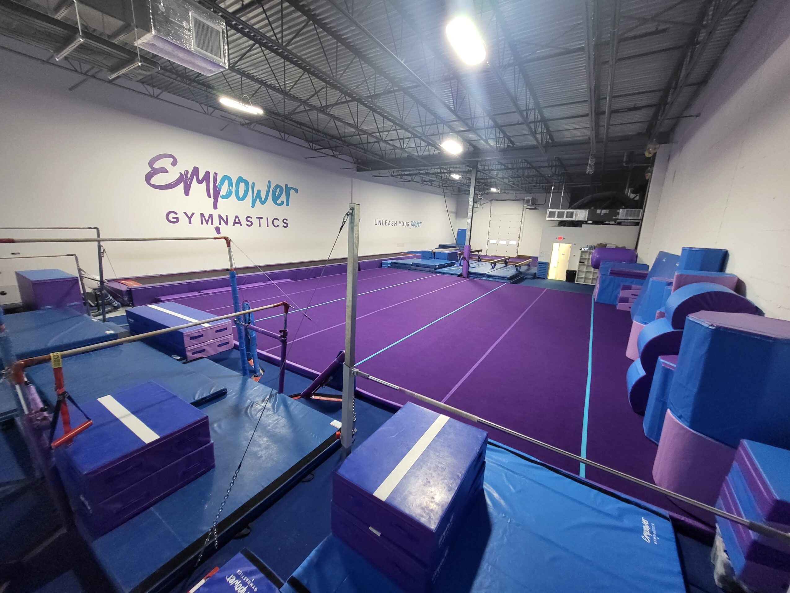 Gymnastics training area with purple mats, balance beams, and foam blocks, inside a spacious gym with high ceilings.