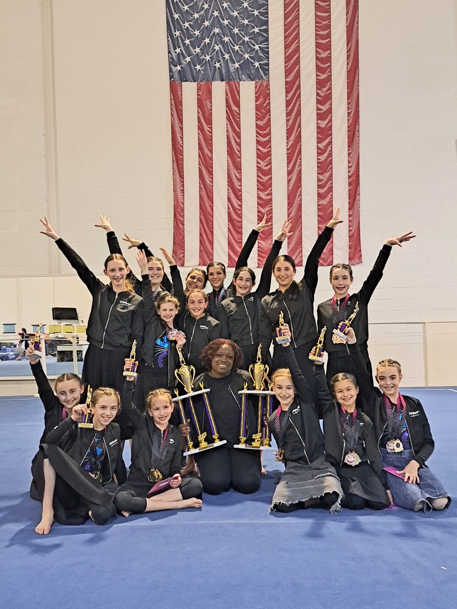 Group of young gymnasts celebrating with trophies in front of an American flag, indoors, smiling, raising arms, some kneeling, some standing.