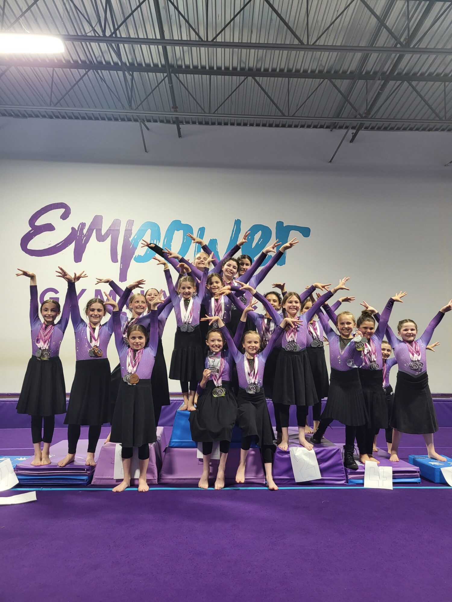 Group of women in purple and black outfits celebrating on stage with raised hands, purple carpet, and a white wall background.