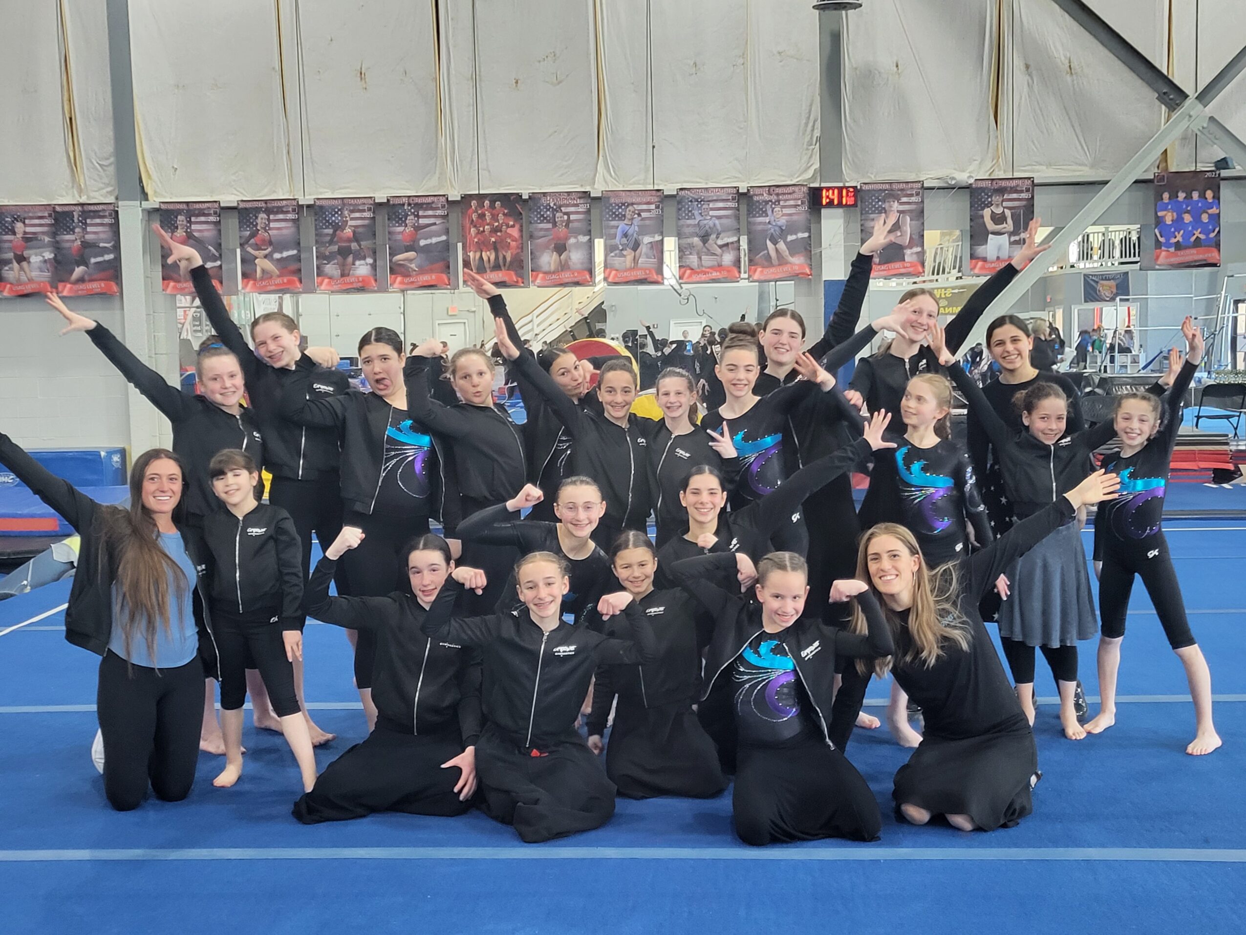 Group of young figure skaters in black costumes and jackets, posing on ice rink with arms raised, indoor arena background.