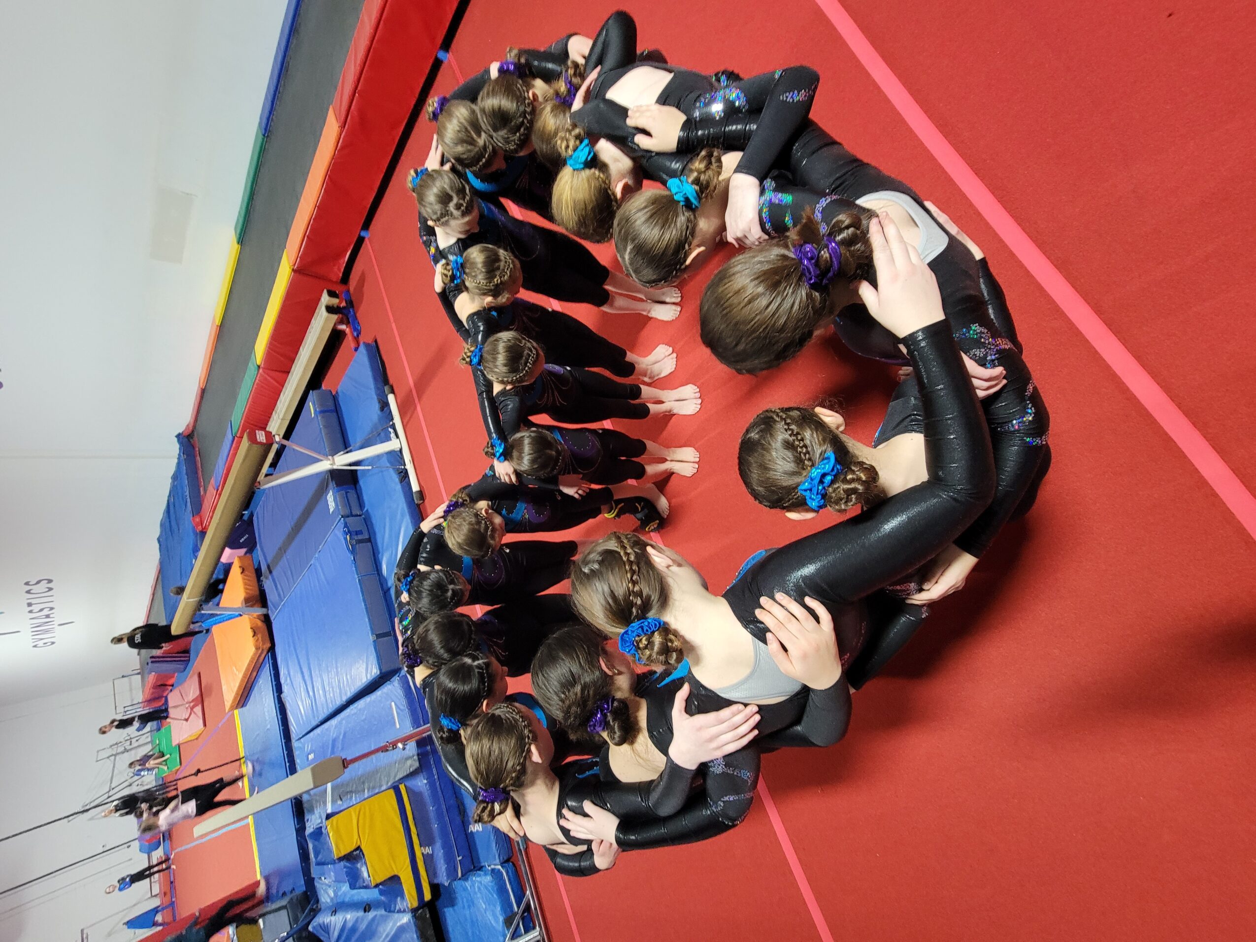 Group of young gymnasts in black leotards sitting in a circle on a red gym floor, listening to their coach.