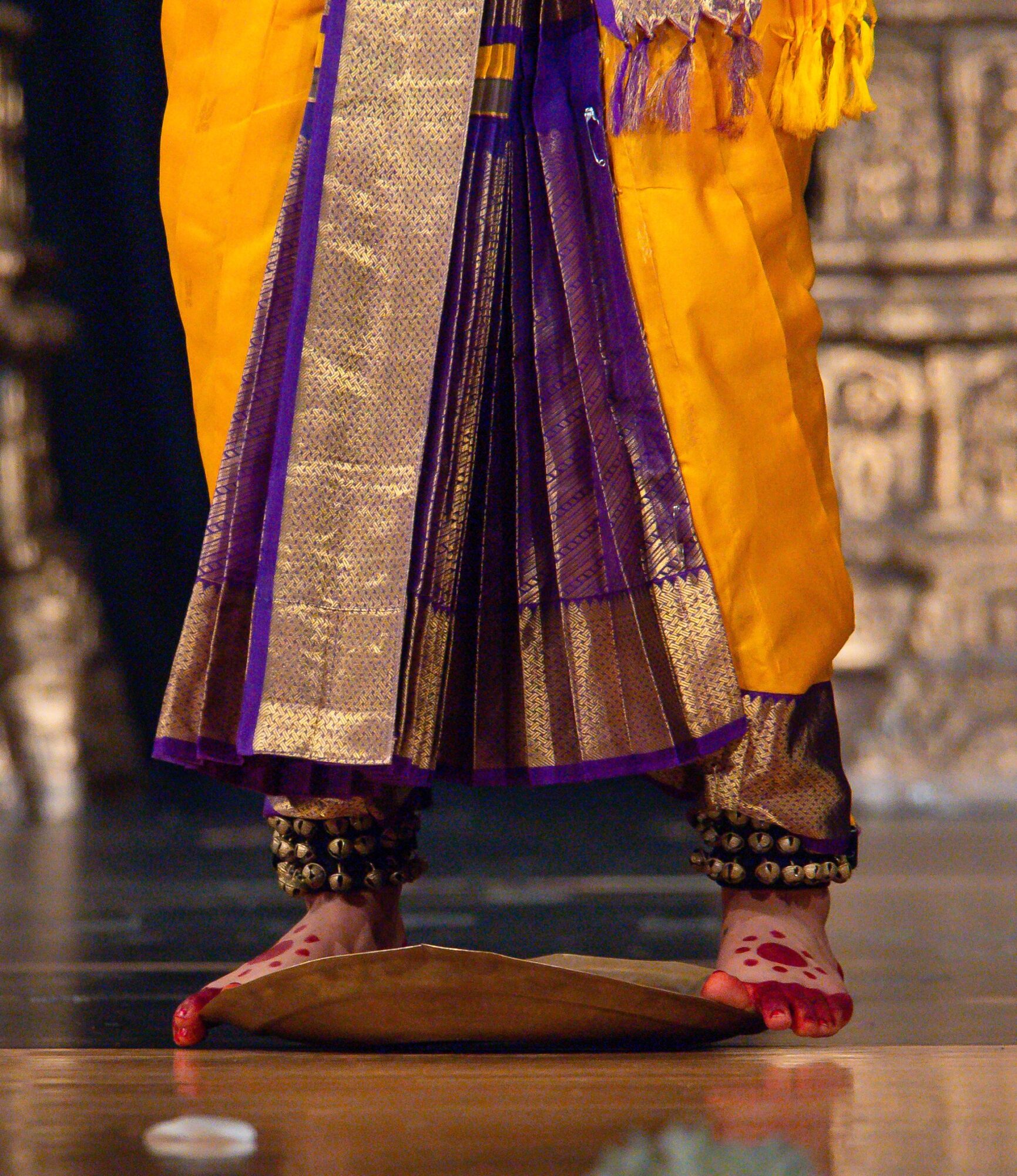 Person standing on a wooden floor wearing traditional attire with colorful fabric, jewelry, and red footwear, with a decorative background.