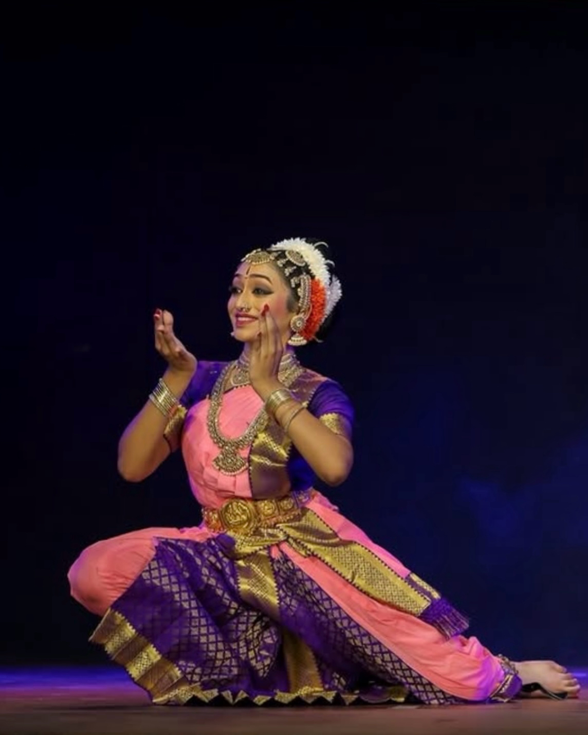 Performer in traditional costume dancing on stage with dark background, smiling, wearing jewelry and floral headpiece.