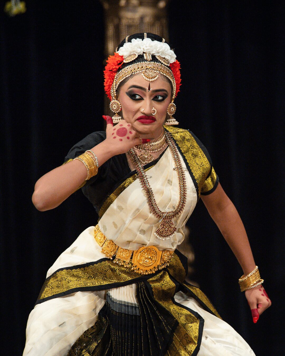 Performer in traditional costume with elaborate jewelry and makeup, striking a pose on stage against a dark background.