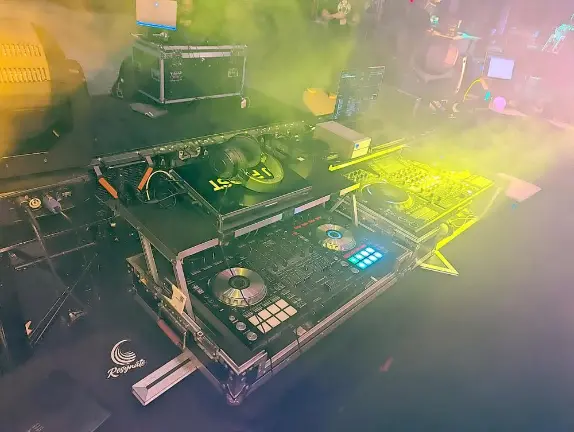 DJ equipment with turntables, mixer, and controllers on a table, surrounded by colorful lights and fog at a party.