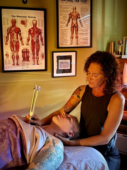 Tattooed woman performs a procedure on a woman lying on a bed in a room with anatomical posters on the wall.