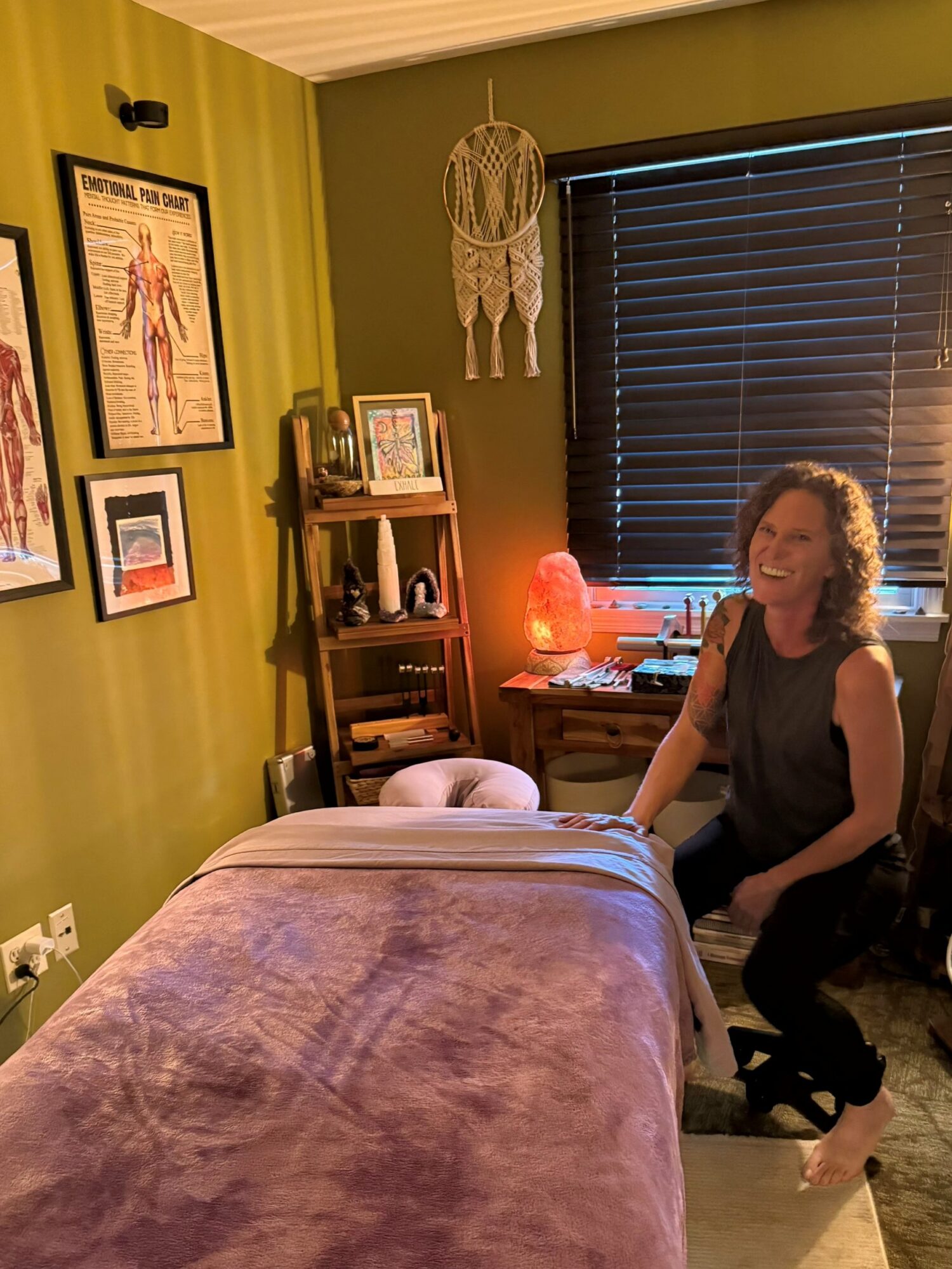 Woman sitting on a chair in a room with green walls, a window with blinds, and a massage table in front.