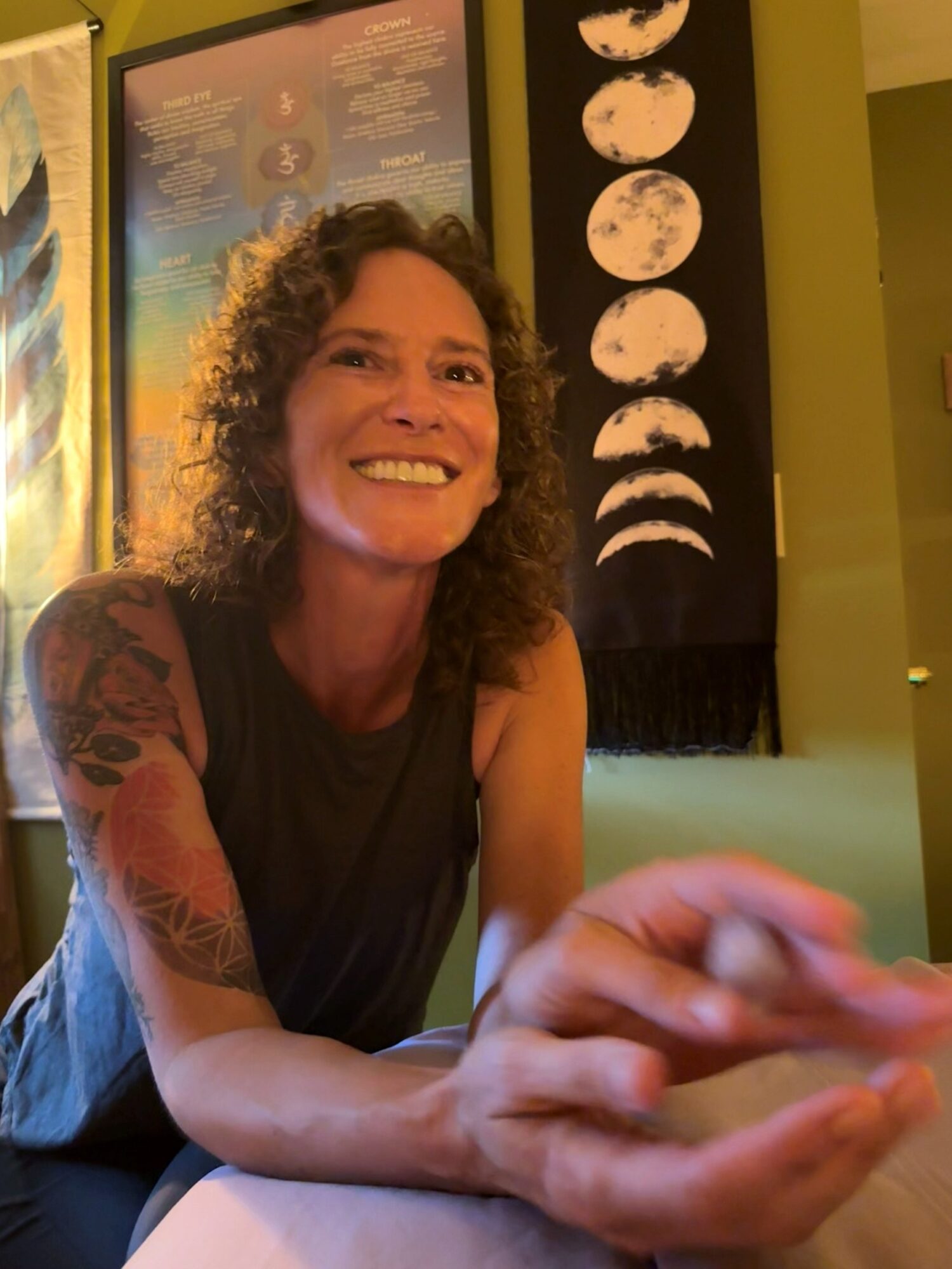 Woman with curly hair smiling, sitting indoors, with a moon phase display behind her, wearing a sleeveless top, arm tattoos visible.