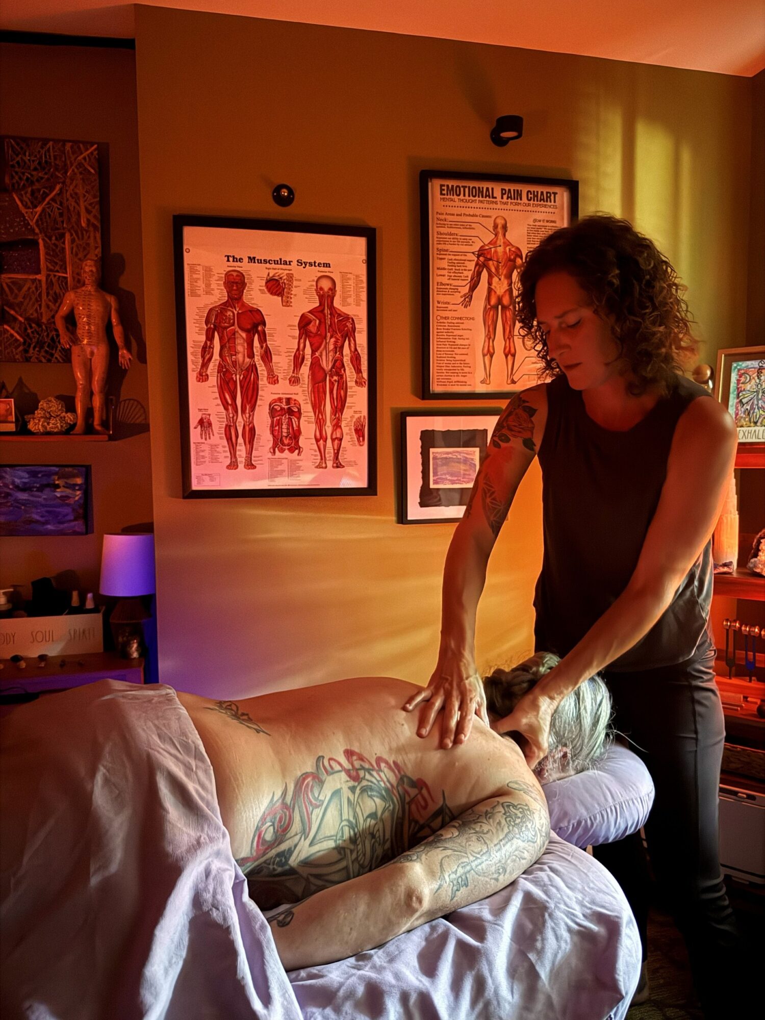 Woman giving massage to person lying face down on massage table in a room with anatomical posters on wall.