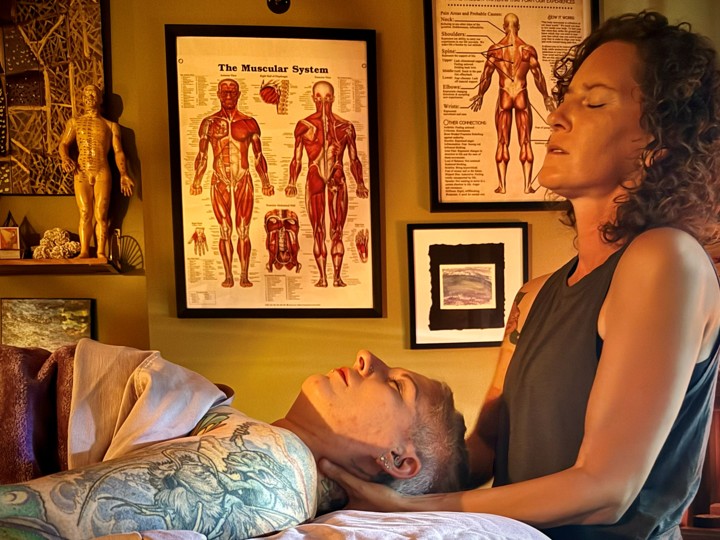 A woman with curly hair looks at a man lying on a table in a room with anatomical posters and decorations.