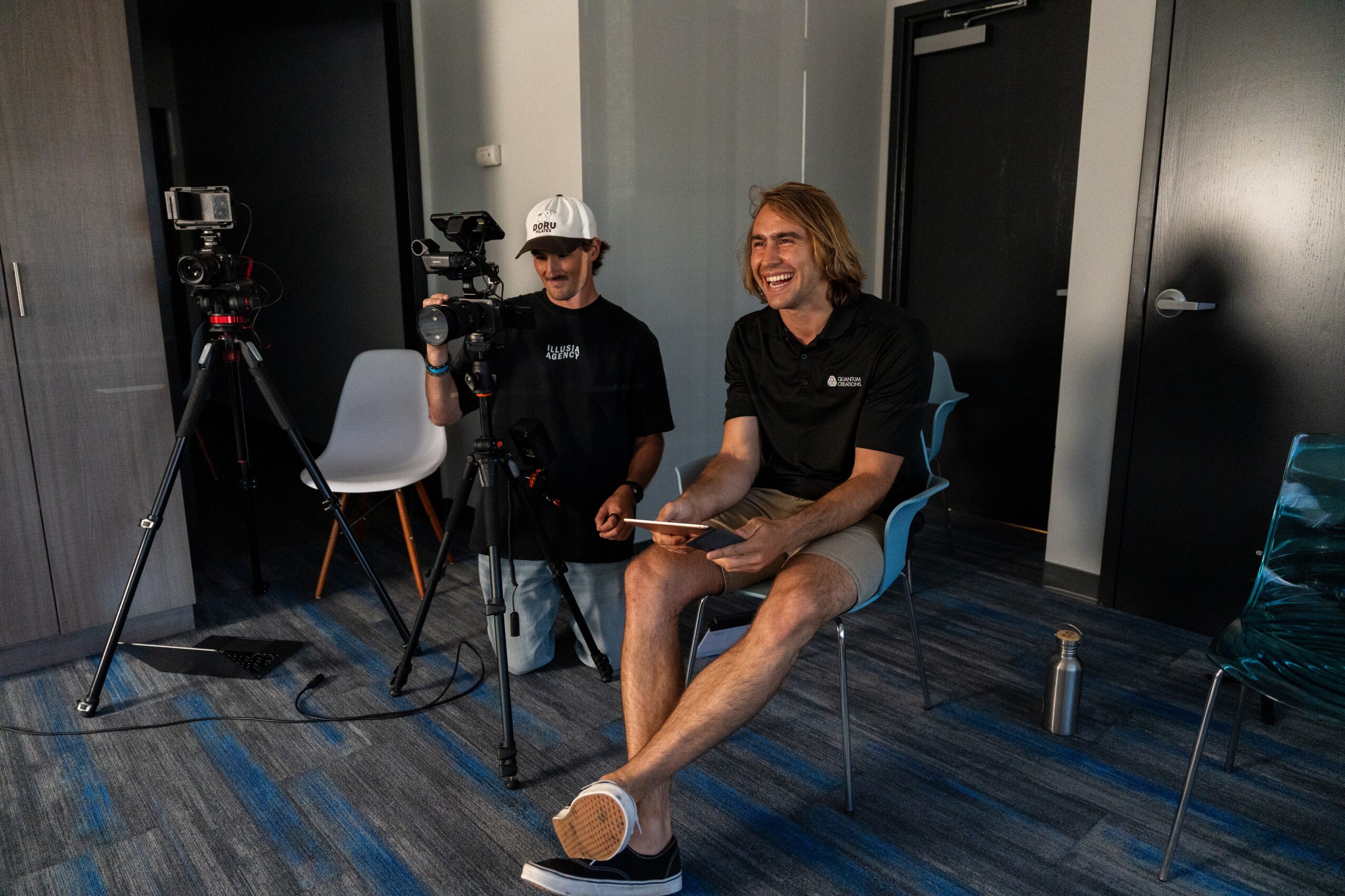 Two people in a room with cameras and equipment, one sitting on a chair and smiling, the other standing nearby.