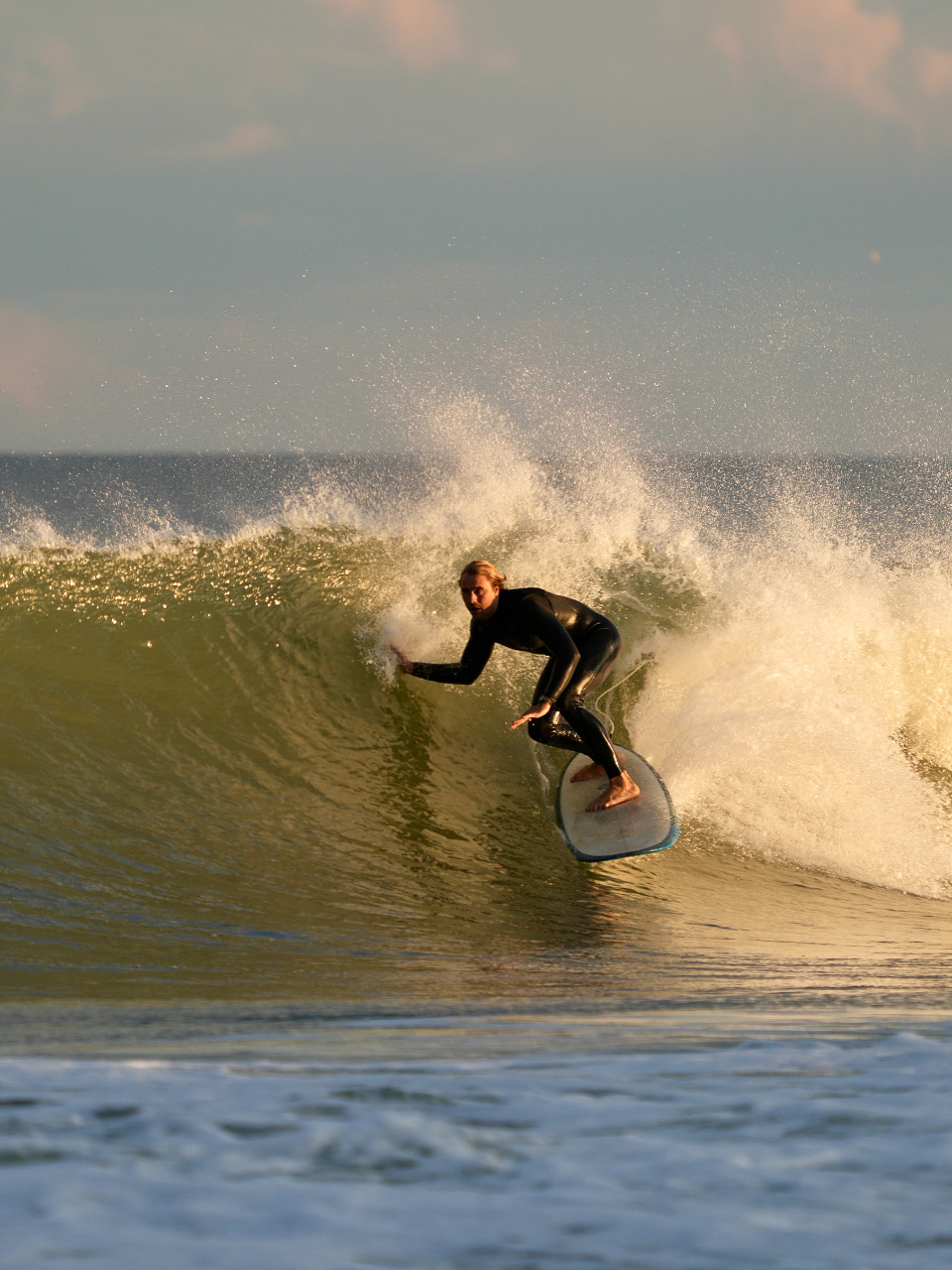 Person surfing on a wave, leaning forward on a surfboard, with spray behind them, under a cloudy sky.