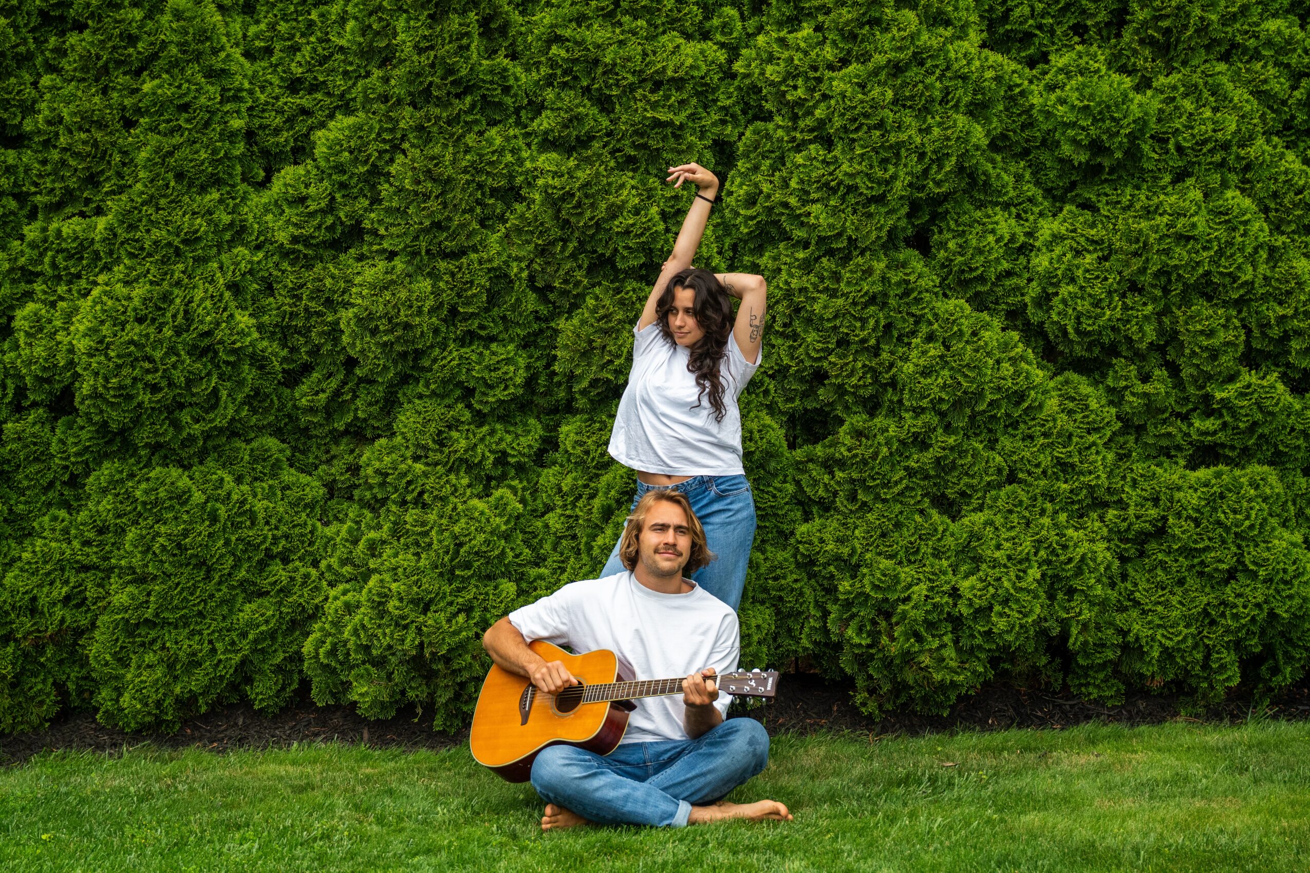 Two people outdoors, one sitting on grass playing guitar, the other standing with arms raised, in front of green bushes.