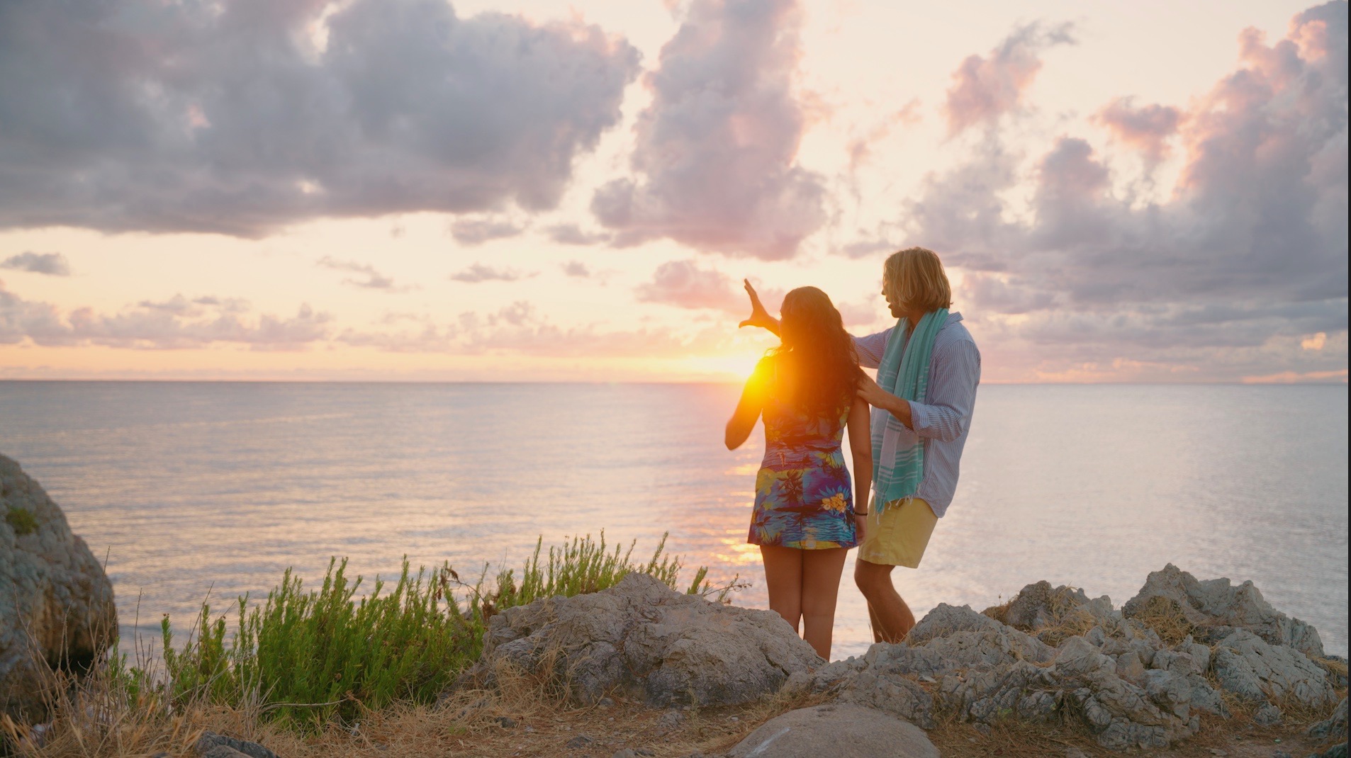 Two people stand on rocks by the water during sunset, with clouds and the ocean in the background.
