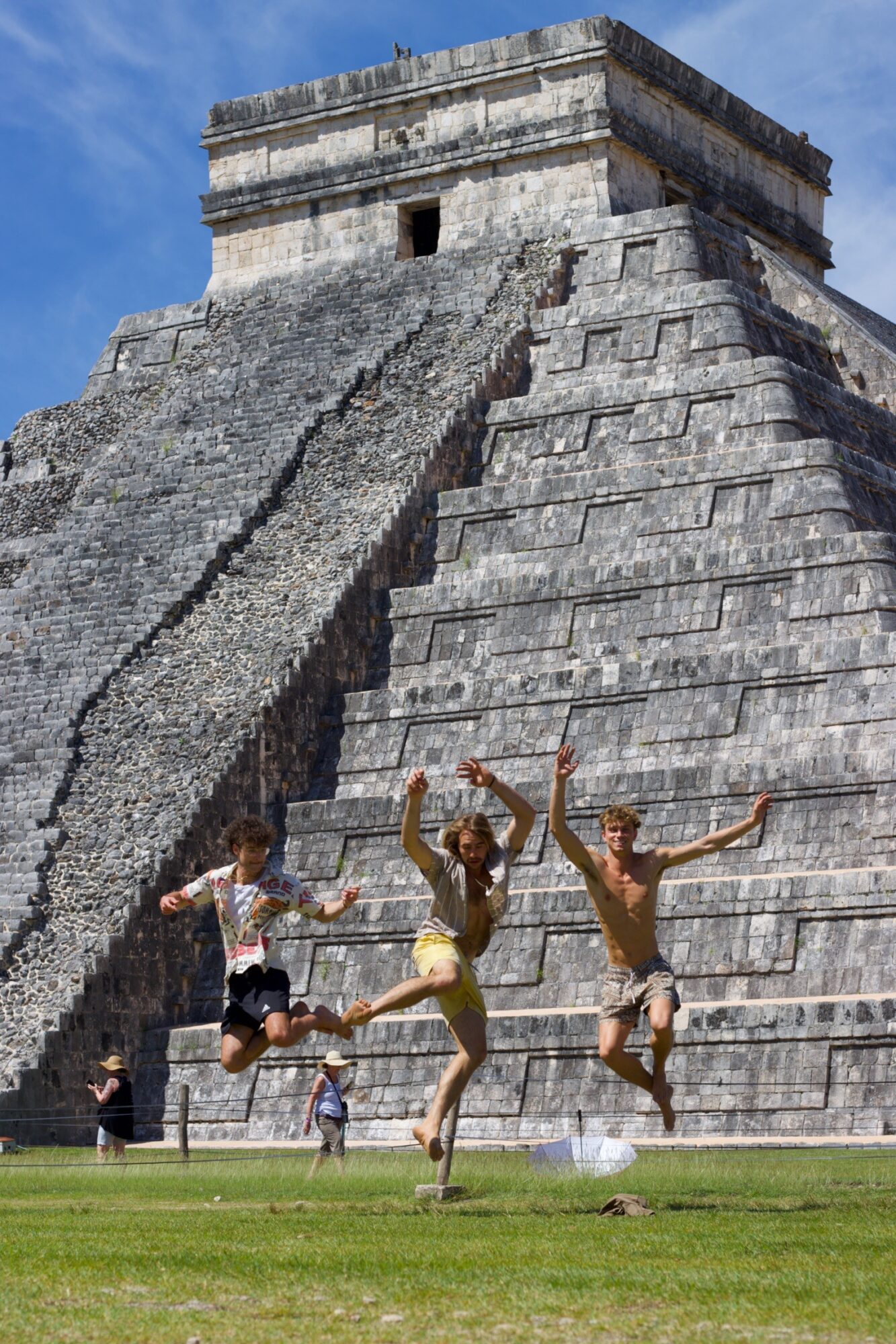 Three children jumping in front of a large ancient stone pyramid with steps, under a blue sky.