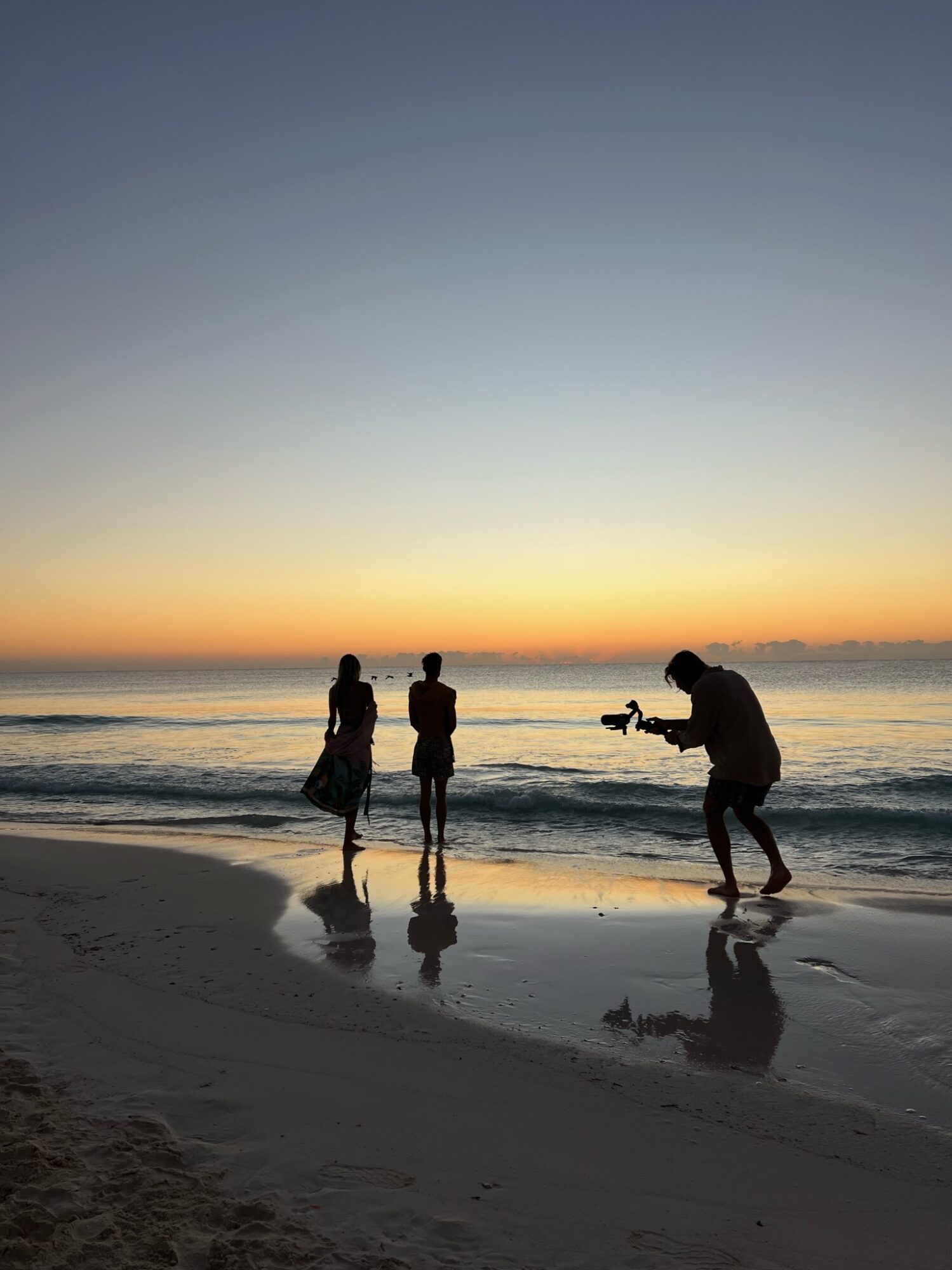 Three people on a beach at sunset, two standing and one taking a photo, with reflections on wet sand.