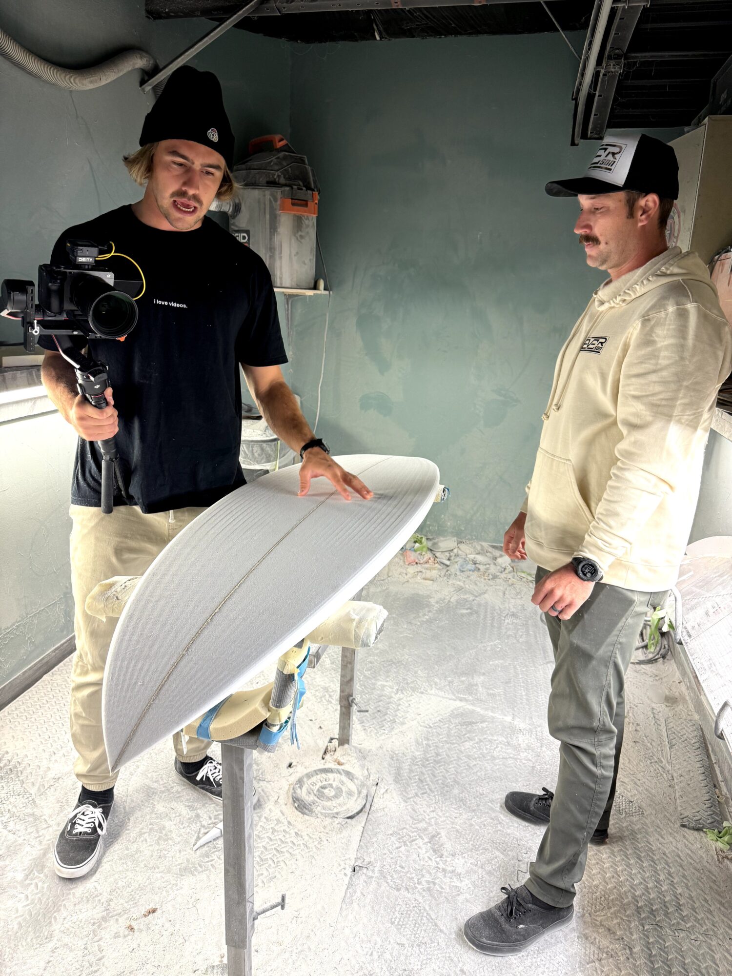 Two men in casual and work attire stand indoors near a surfboard on a stand, with a camera operator filming them.