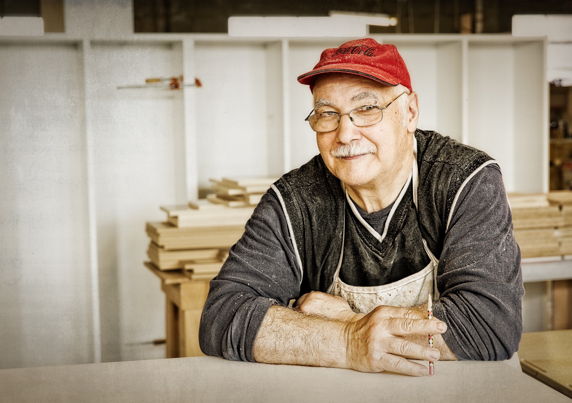 Elderly man with glasses and a red cap, smiling, sitting at a workbench in a woodworking shop.