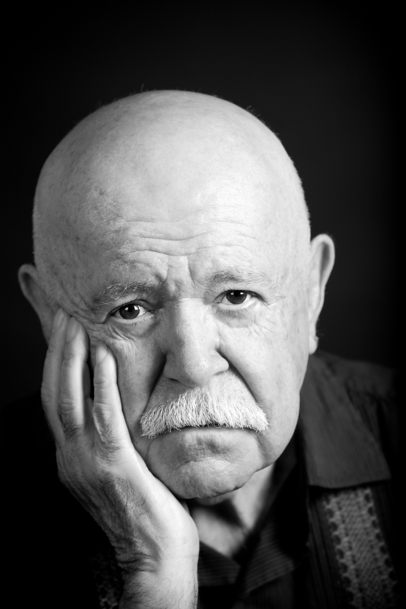 Elderly man with a bald head and mustache, resting his face on his hand, looking thoughtful, in black and white.