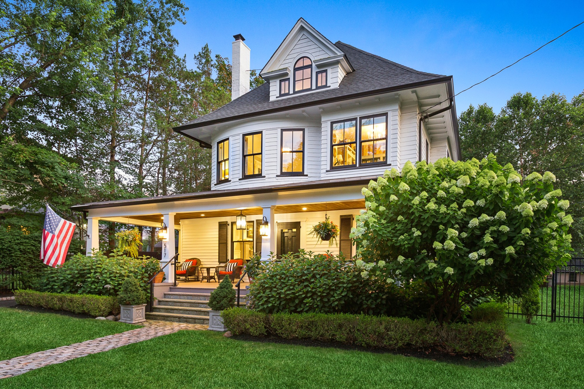 Large white house with a porch, surrounded by green bushes and trees, under a blue sky.