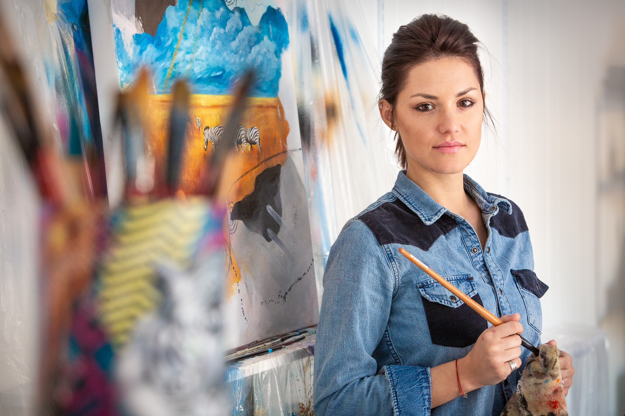 Young woman holding a paintbrush and palette, standing in front of an abstract painting, with art supplies in foreground.