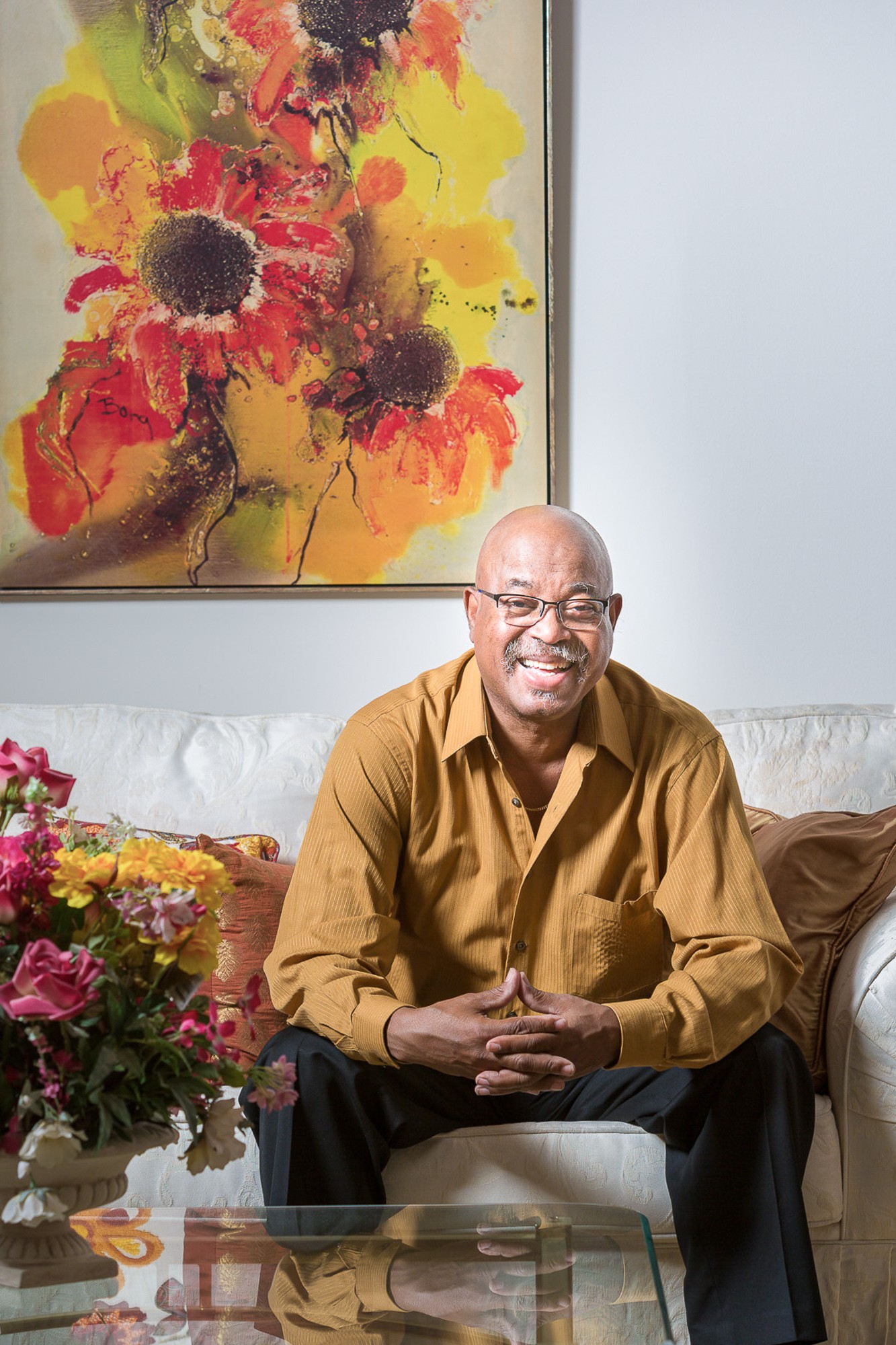 Smiling man with glasses in a brown shirt sitting on a sofa with flowers and a colorful painting behind him.
