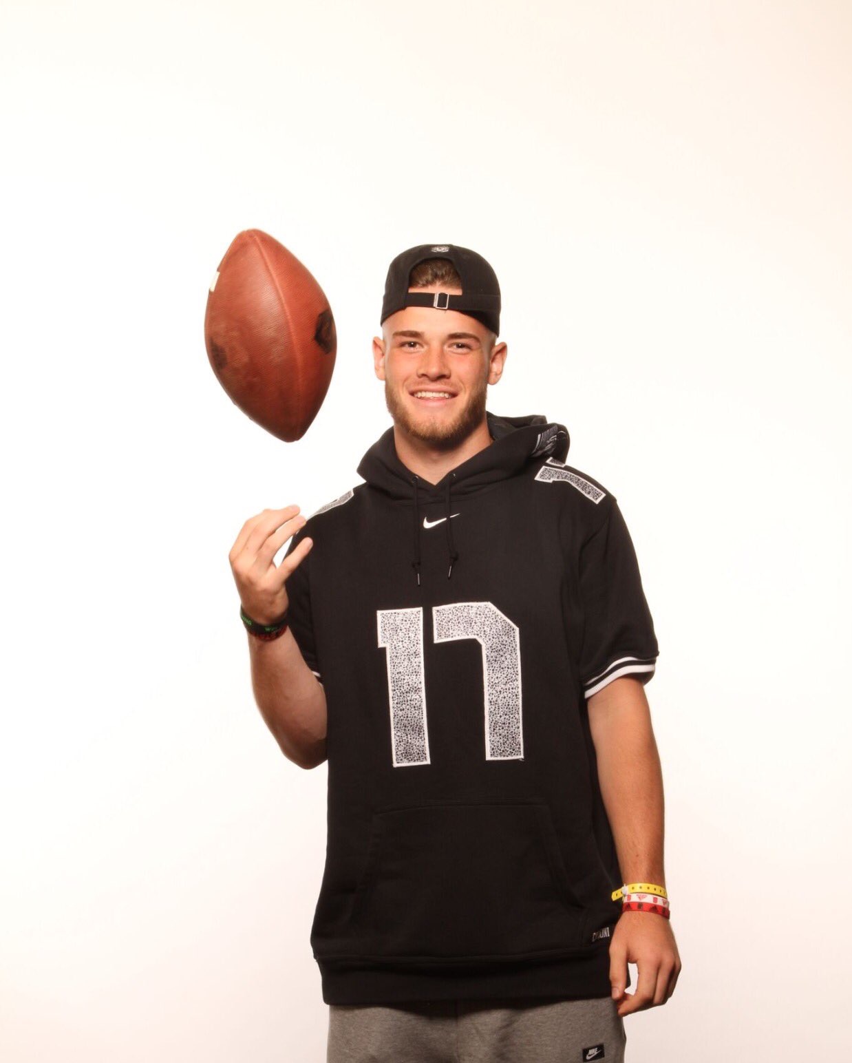 Young man in black football jersey with number 17, smiling, wearing a cap backward, holding a football, standing against a white background.