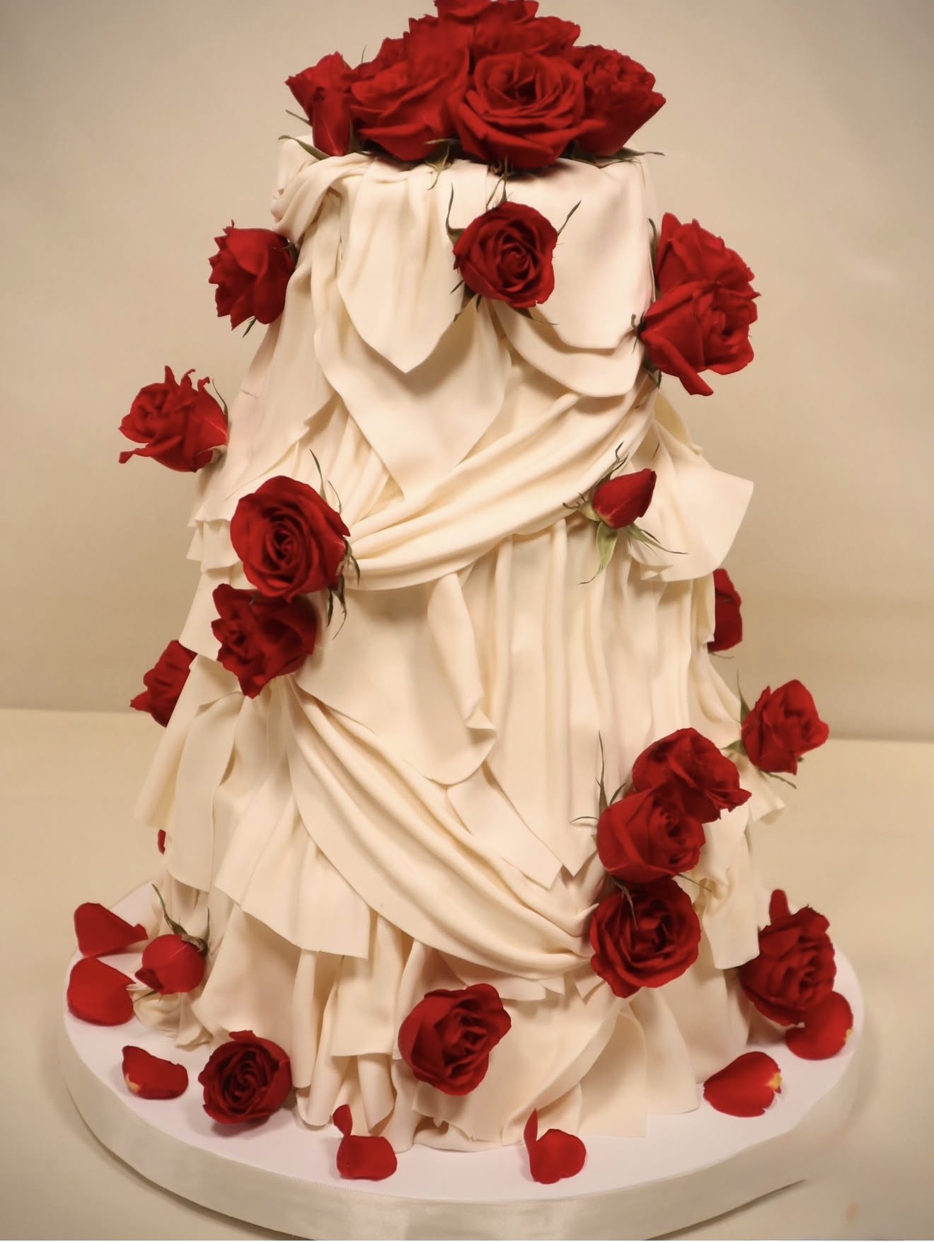 Cake decorated with white fabric and red roses, some roses on the cake and others scattered around base.