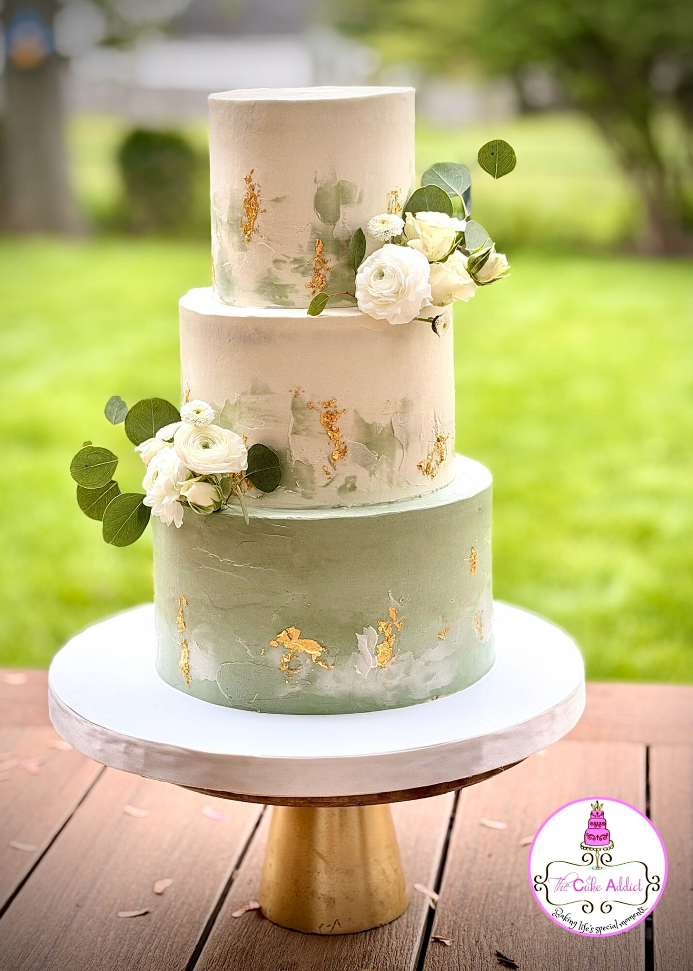 Three-tiered cake with white, light blue, and beige colors, decorated with white flowers and green leaves, outdoors on a table.