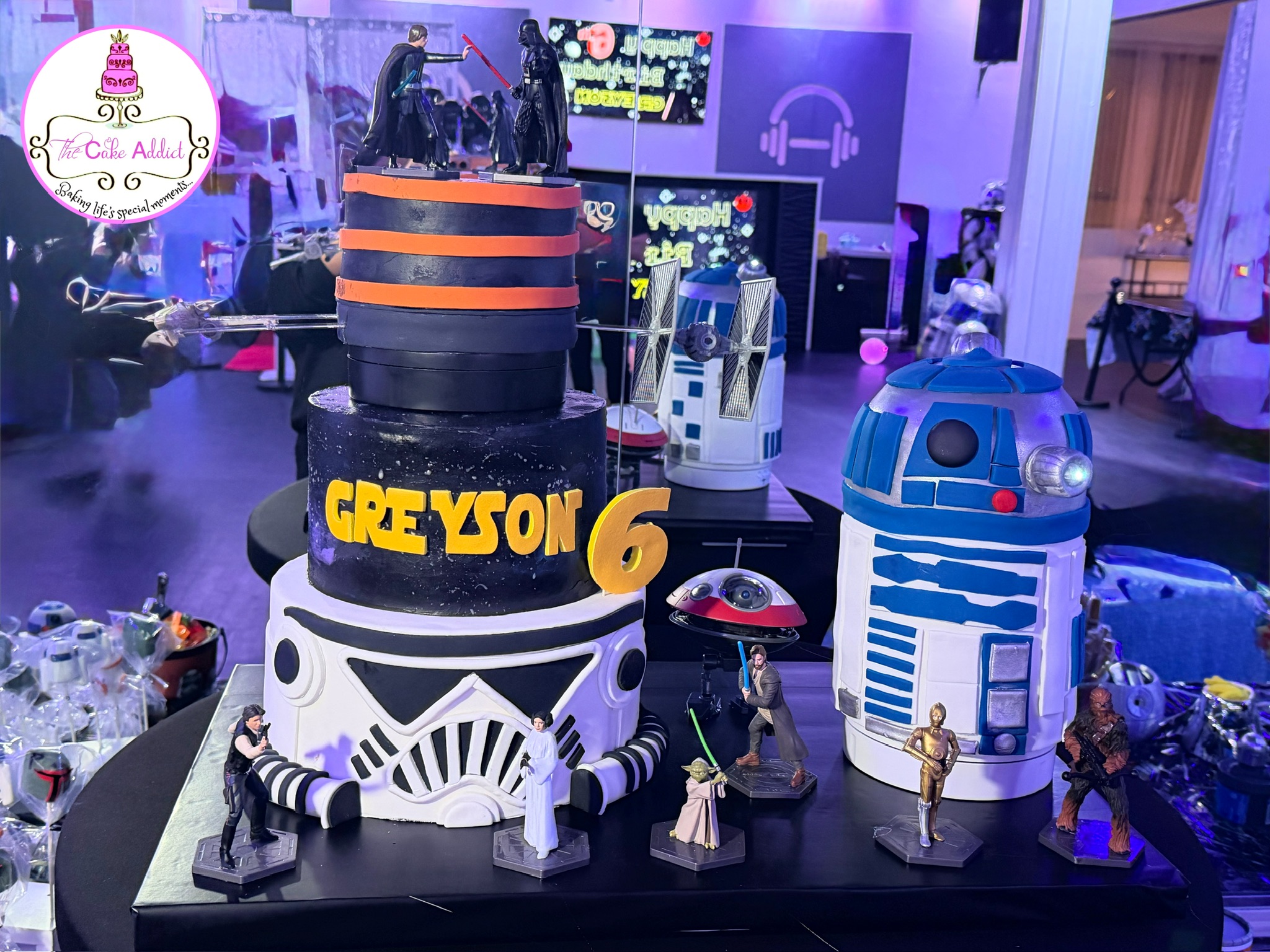 Star Wars-themed cake with R2-D2 and a large black and orange structure, surrounded by small figures and decorations, in a party setting.