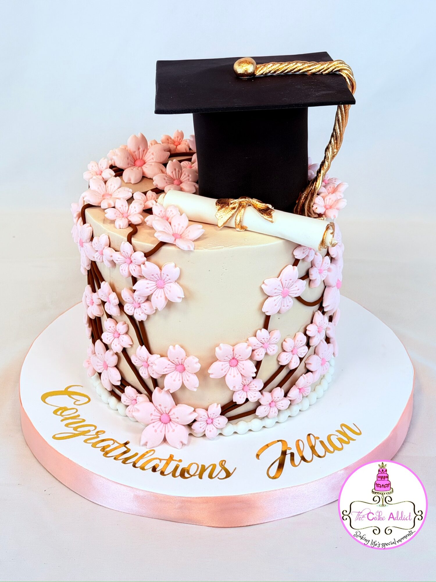 Graduation cake with pink floral decorations, a black mortarboard cap, and a diploma, on a white base with gold writing.