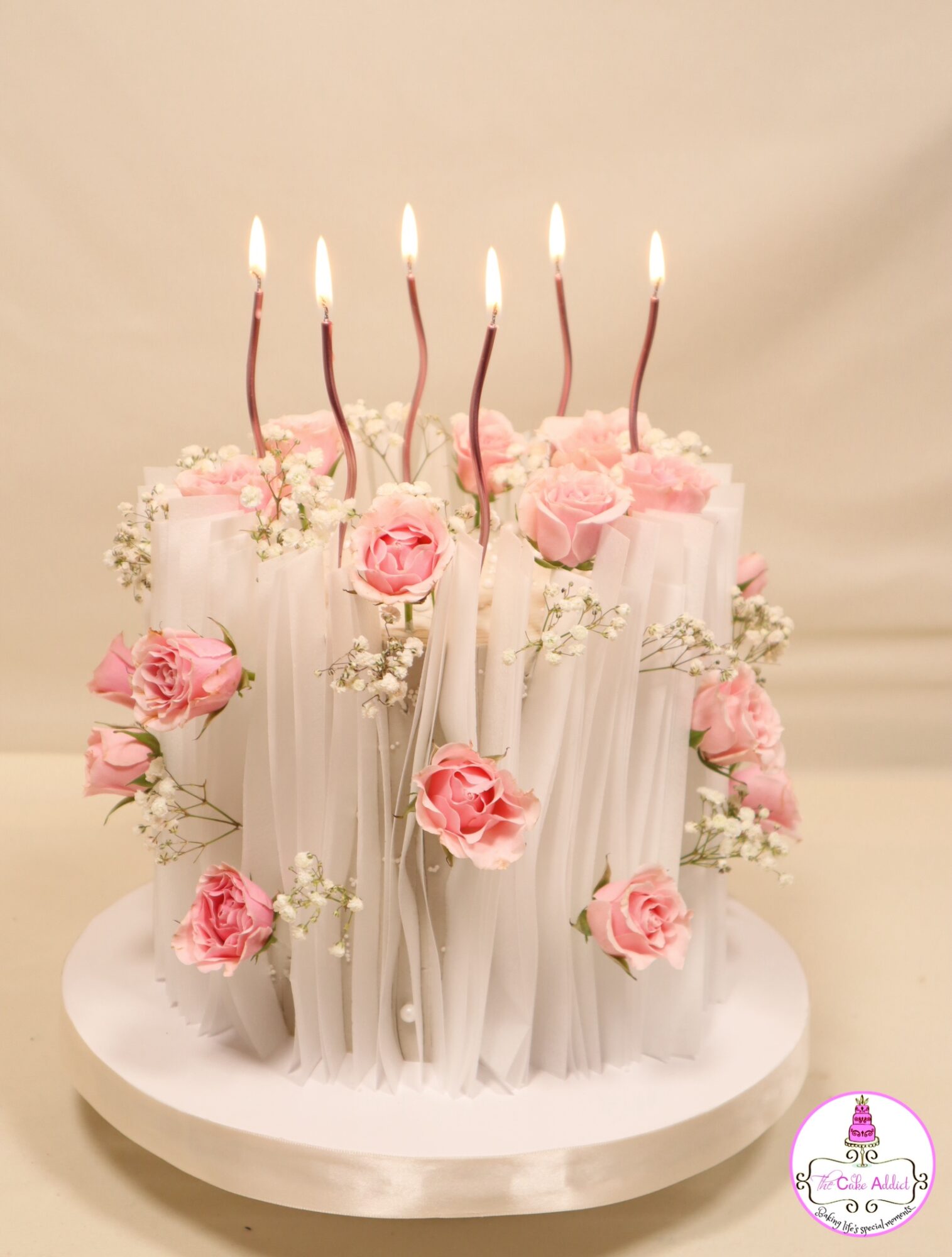 Cake decorated with pink roses, white flowers, and lit candles on top, placed on a white round base.
