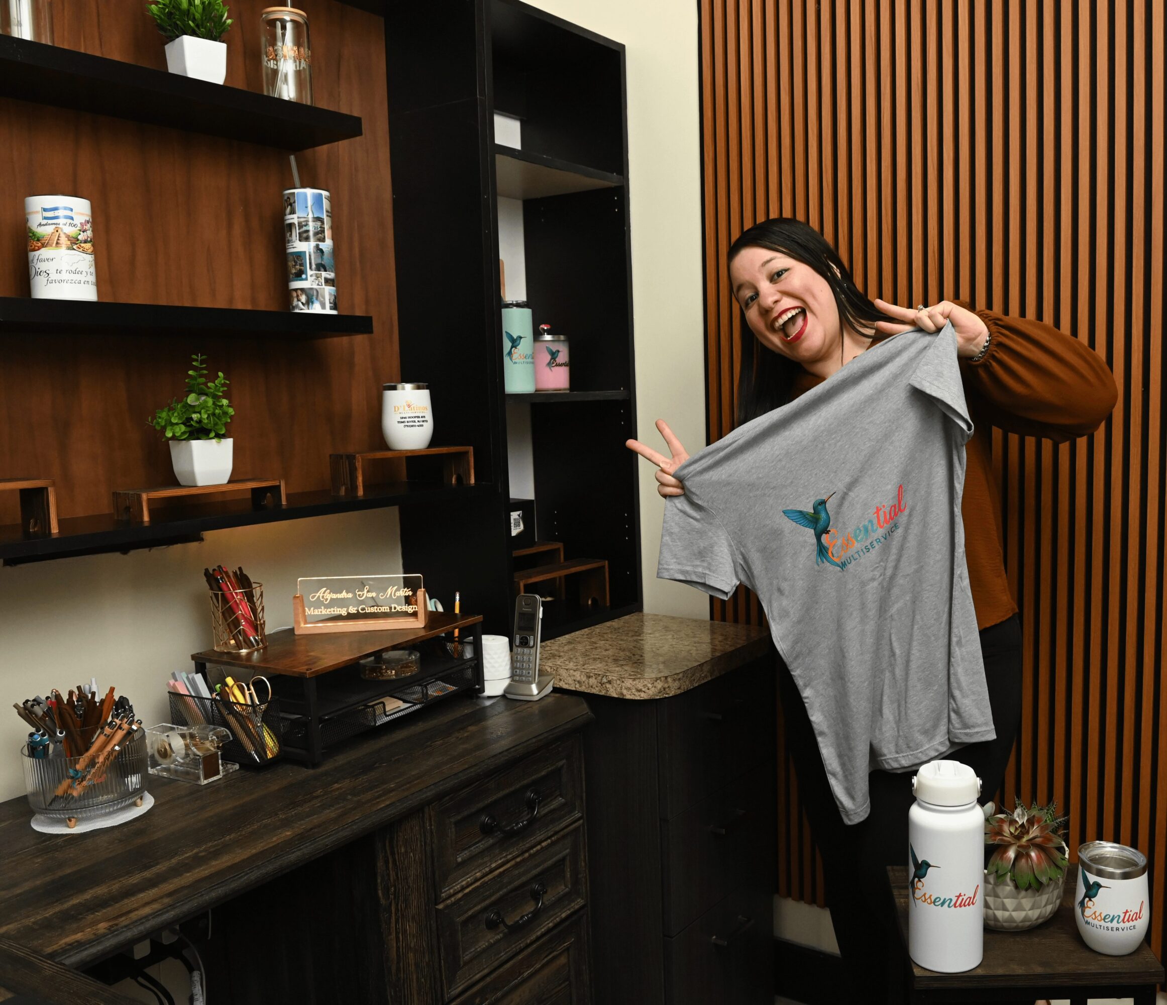 Woman smiling and holding a gray T-shirt with a colorful bird design in a modern kitchen.