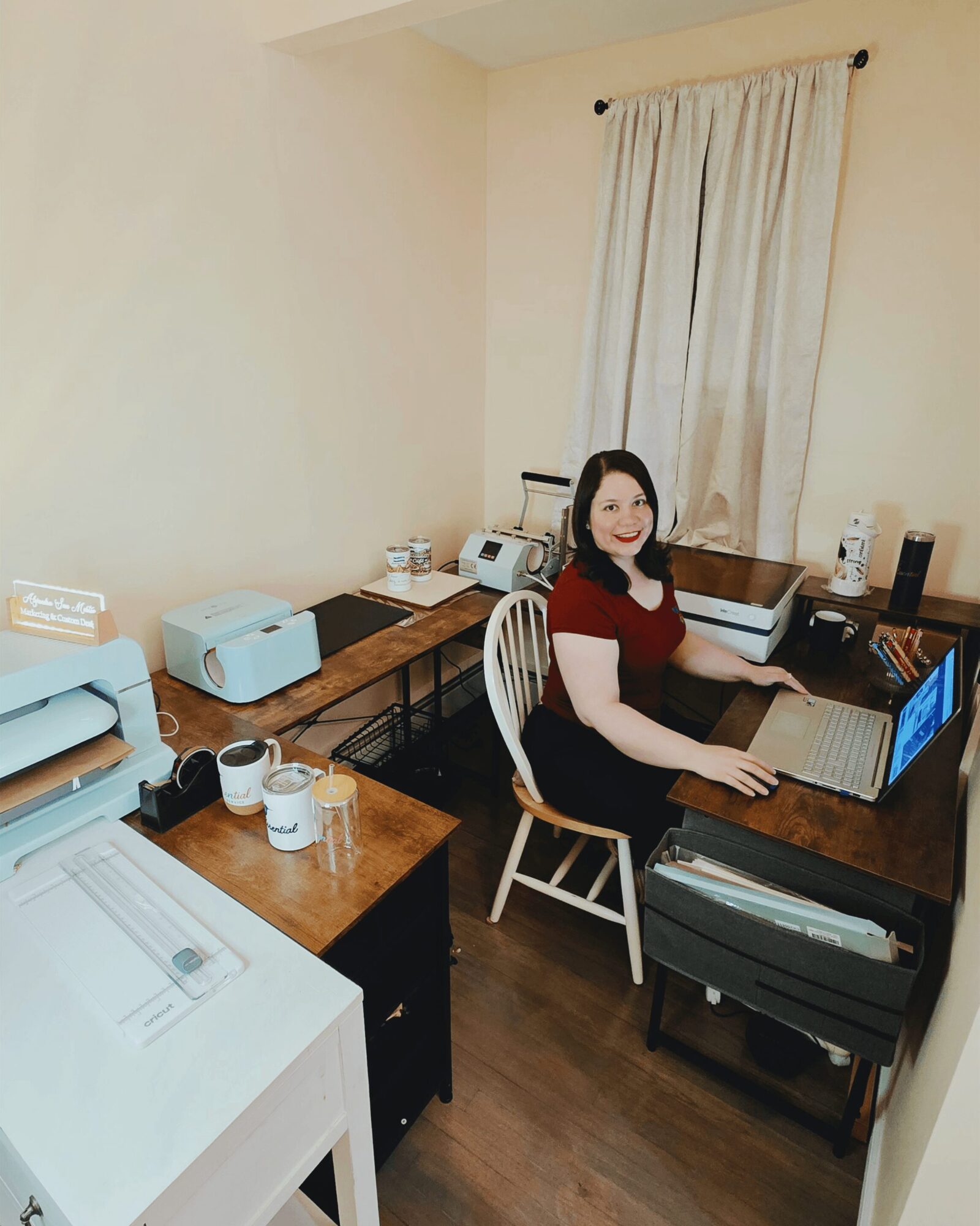 Woman sitting at a desk with a laptop, surrounded by office equipment and supplies, in a room with a window and curtain.