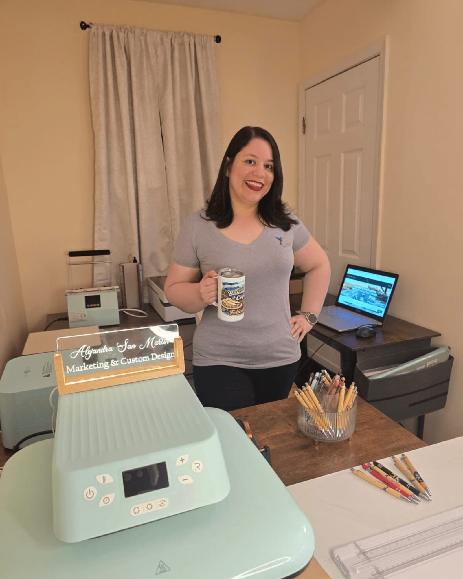 Woman holding a mug, smiling, standing in a room with a laptop and printer, beige walls, and a window with curtains.