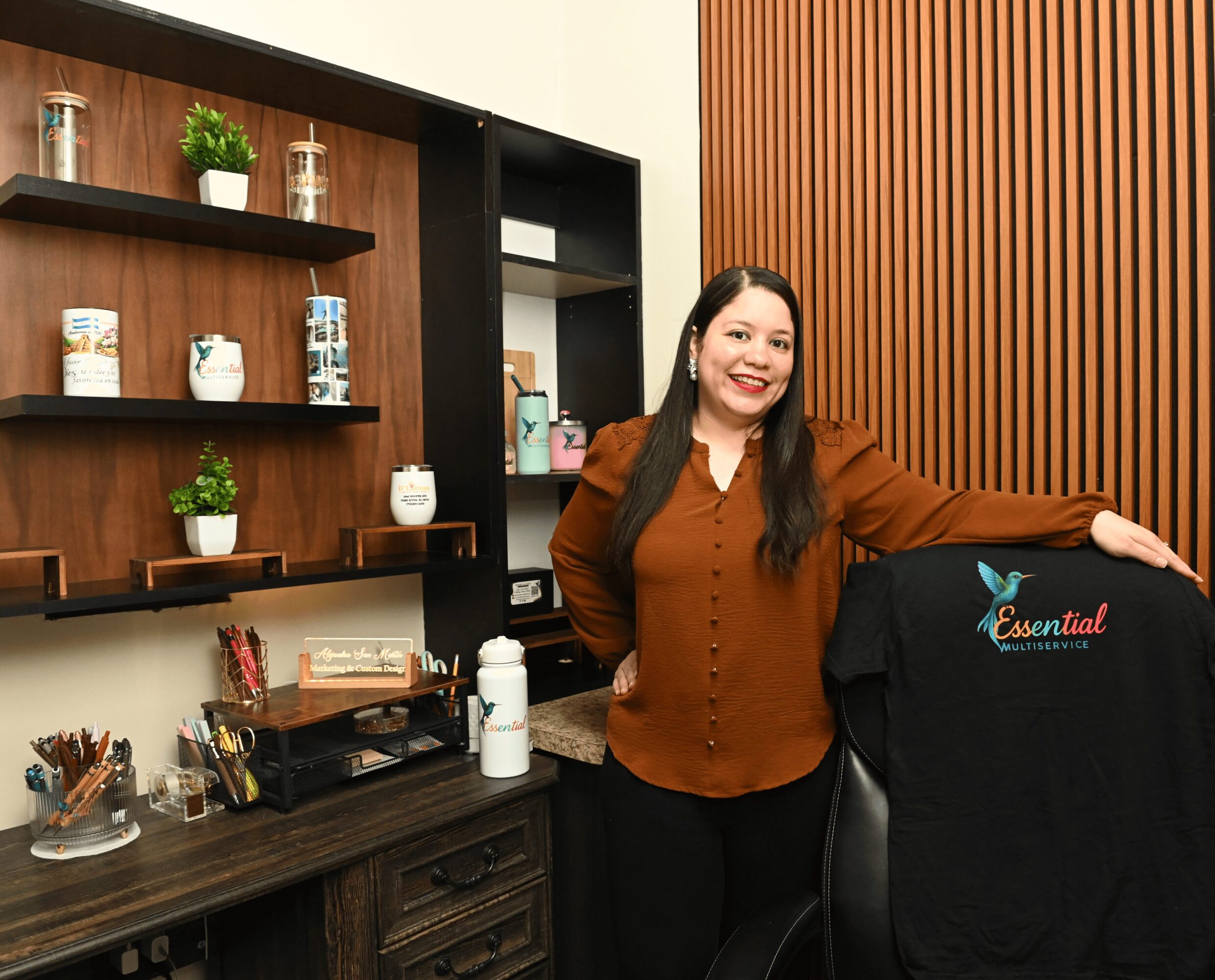 Woman standing in an office or store, smiling, with shelves and a desk behind her.
