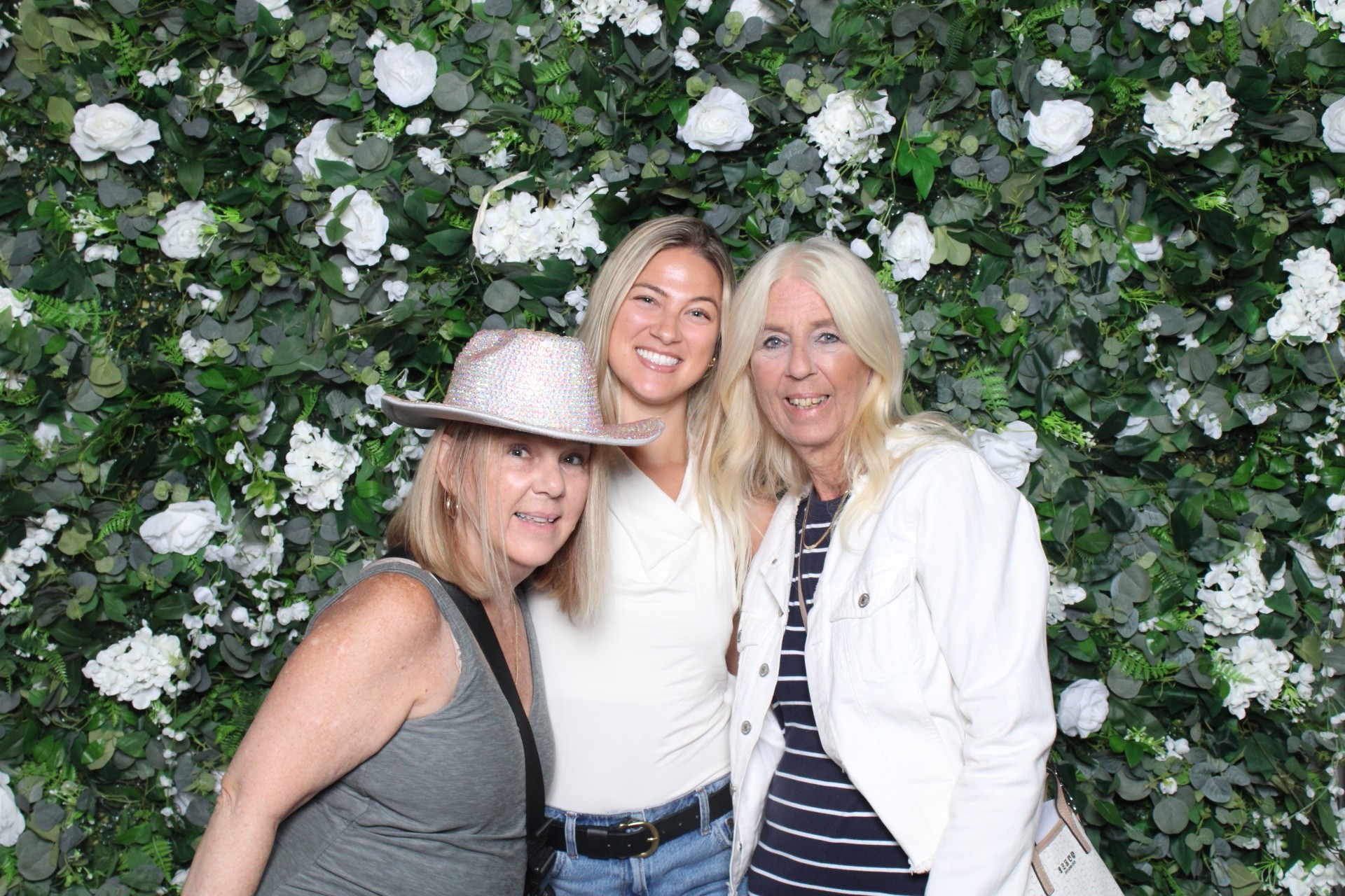 Three women smiling in front of a floral backdrop with white flowers and green leaves.