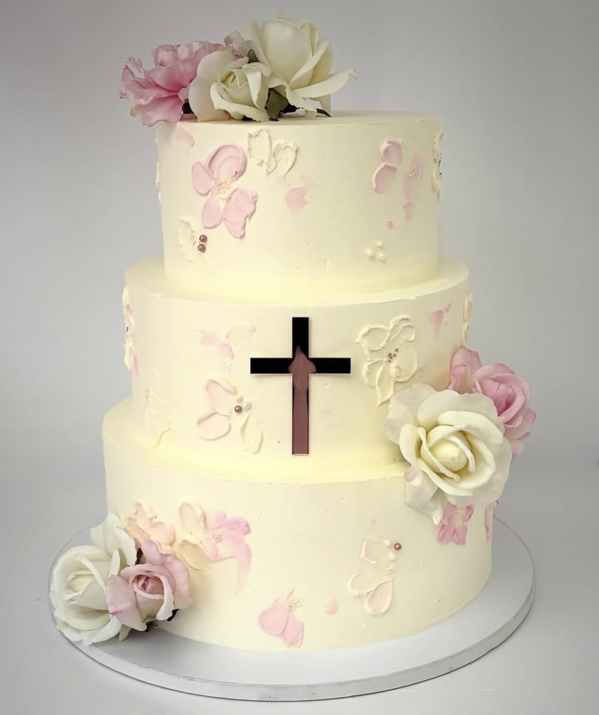 Three-tiered cake decorated with pink and white flowers, featuring a black cross cutout on the middle tier, on a white base.