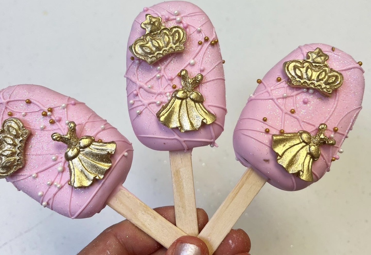 Three pink and purple decorated cake pops with gold crown and dress charms, held by a hand.