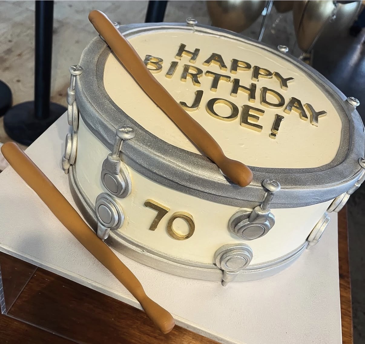 Round birthday cake with drum design, musical notes, and drumsticks, celebrating Joe's 70th birthday, with text on top and side.