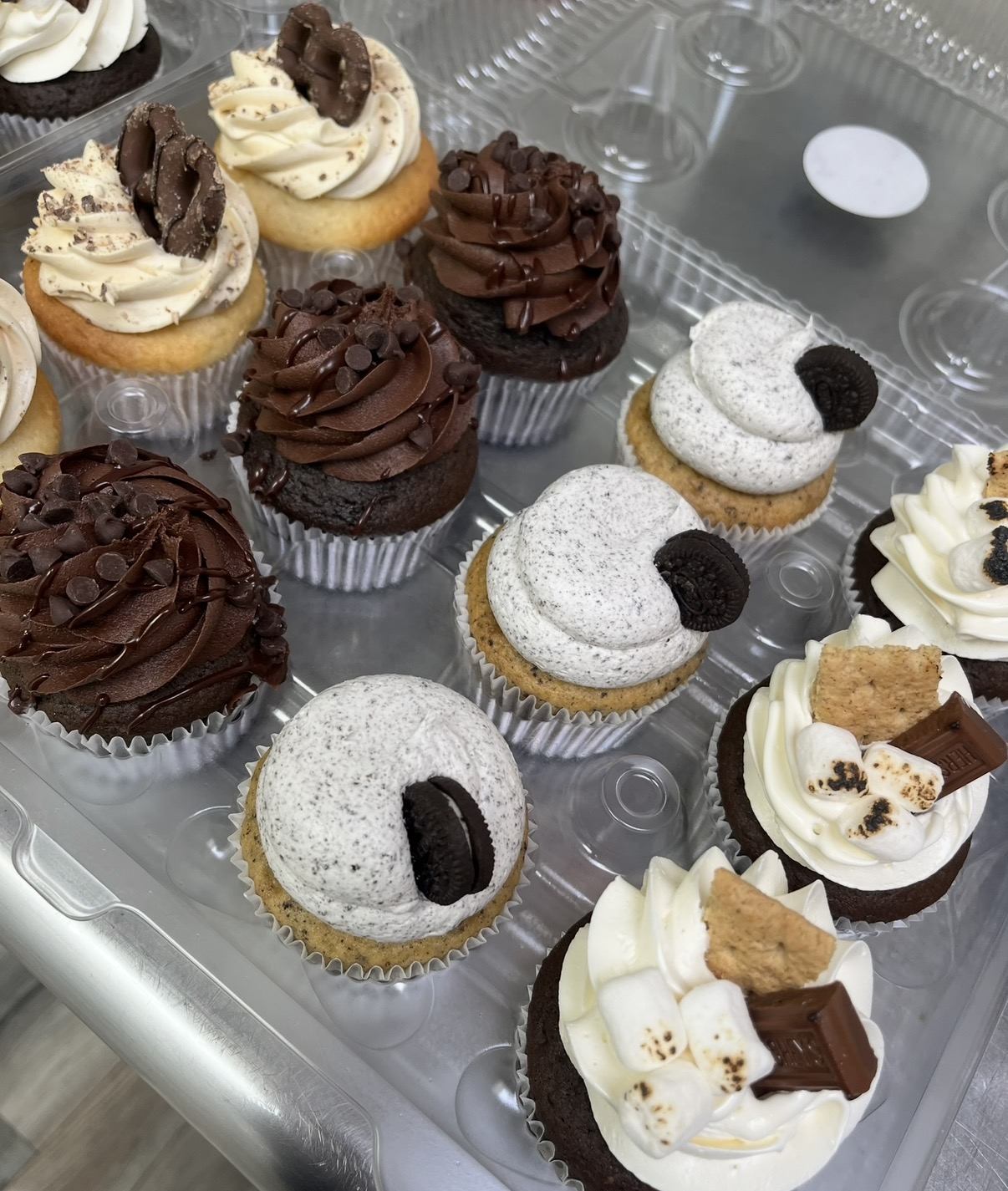 Assorted cupcakes with chocolate, vanilla, and cookies and cream frosting, some topped with cookies or chocolate pieces, on a clear plastic tray.