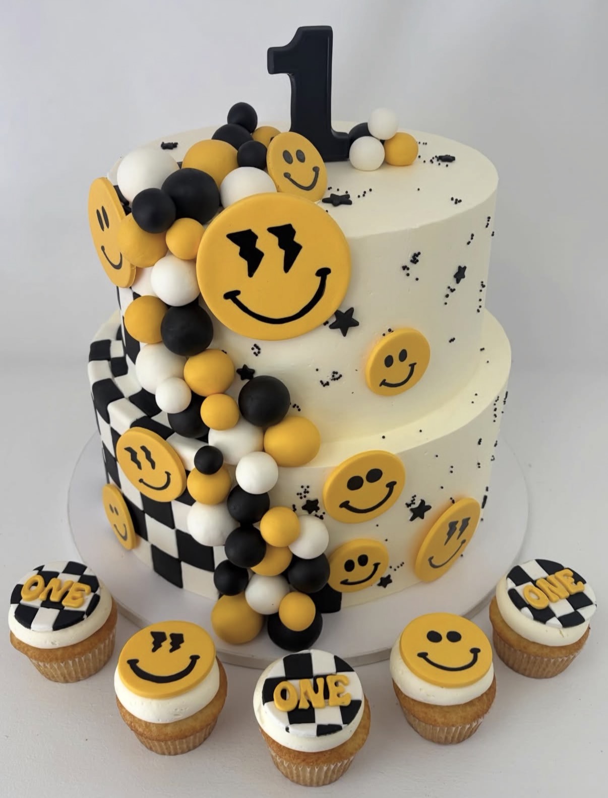 Three-tiered birthday cake decorated with black, white, and yellow smiley faces, checkered patterns, and a large number one topper, surrounded by cupcakes.