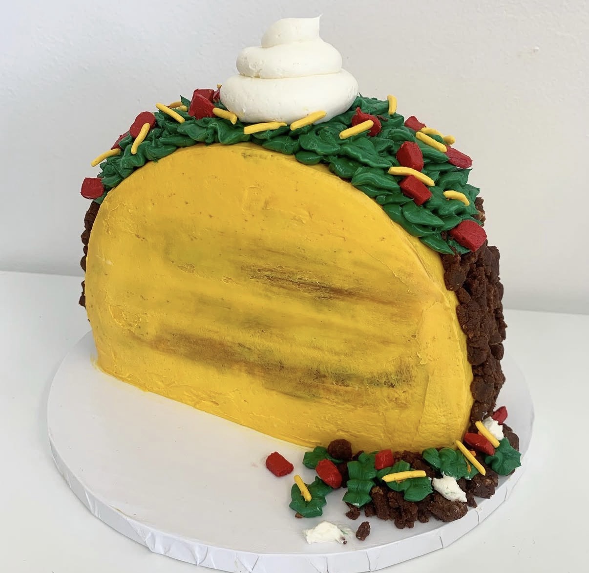 Cake shaped like a taco with yellow shell, green, red, and yellow decorations, topped with white whipped cream.