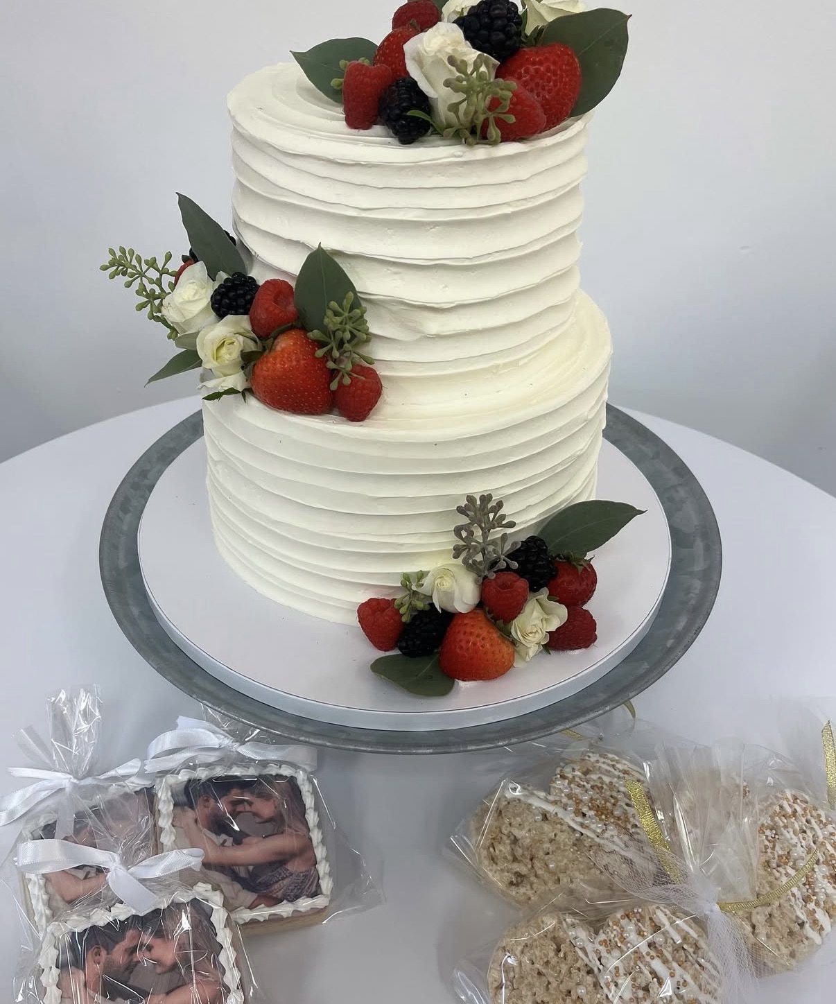 Two-tiered white cake decorated with berries and flowers, on a silver cake stand, with wrapped treats in front.