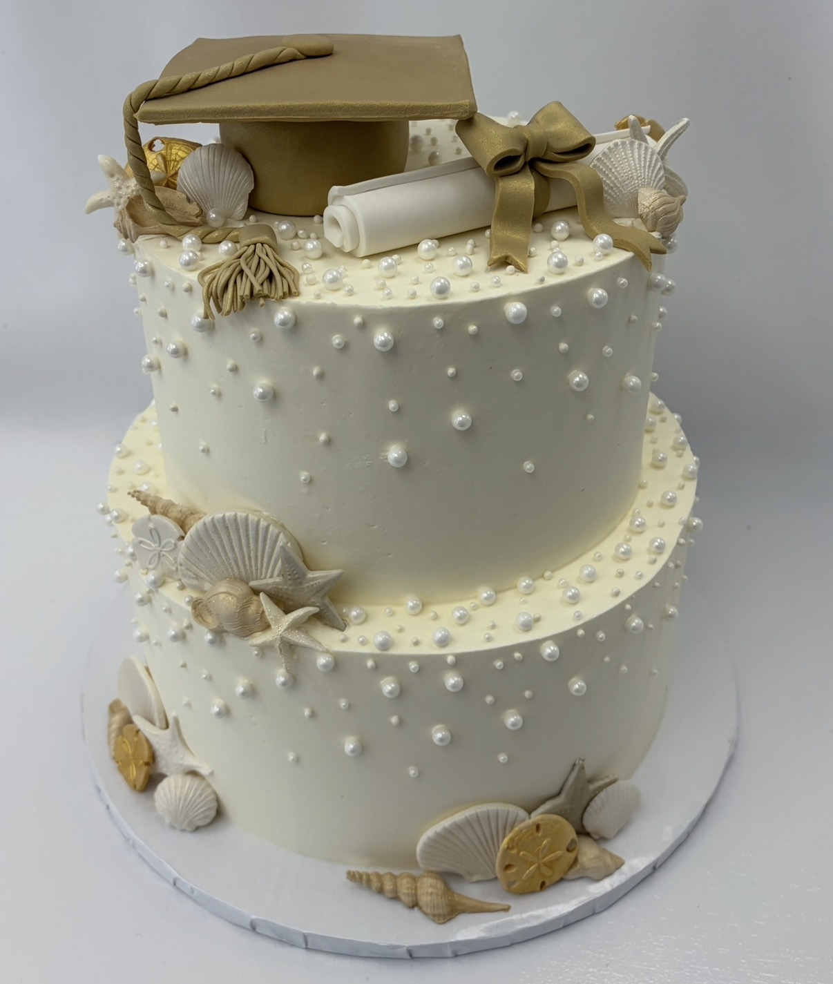 Two-tiered white cake decorated with shells, pearls, and a rolled scroll with a bow on top, with gift boxes and cookies around base.