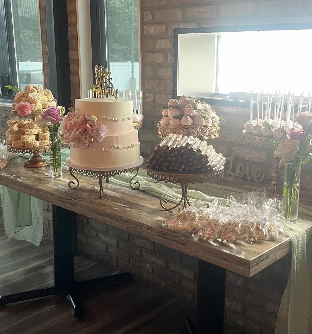 Dessert table with cake, cupcakes, flowers, and treats against brick wall and window, decorated for celebration.